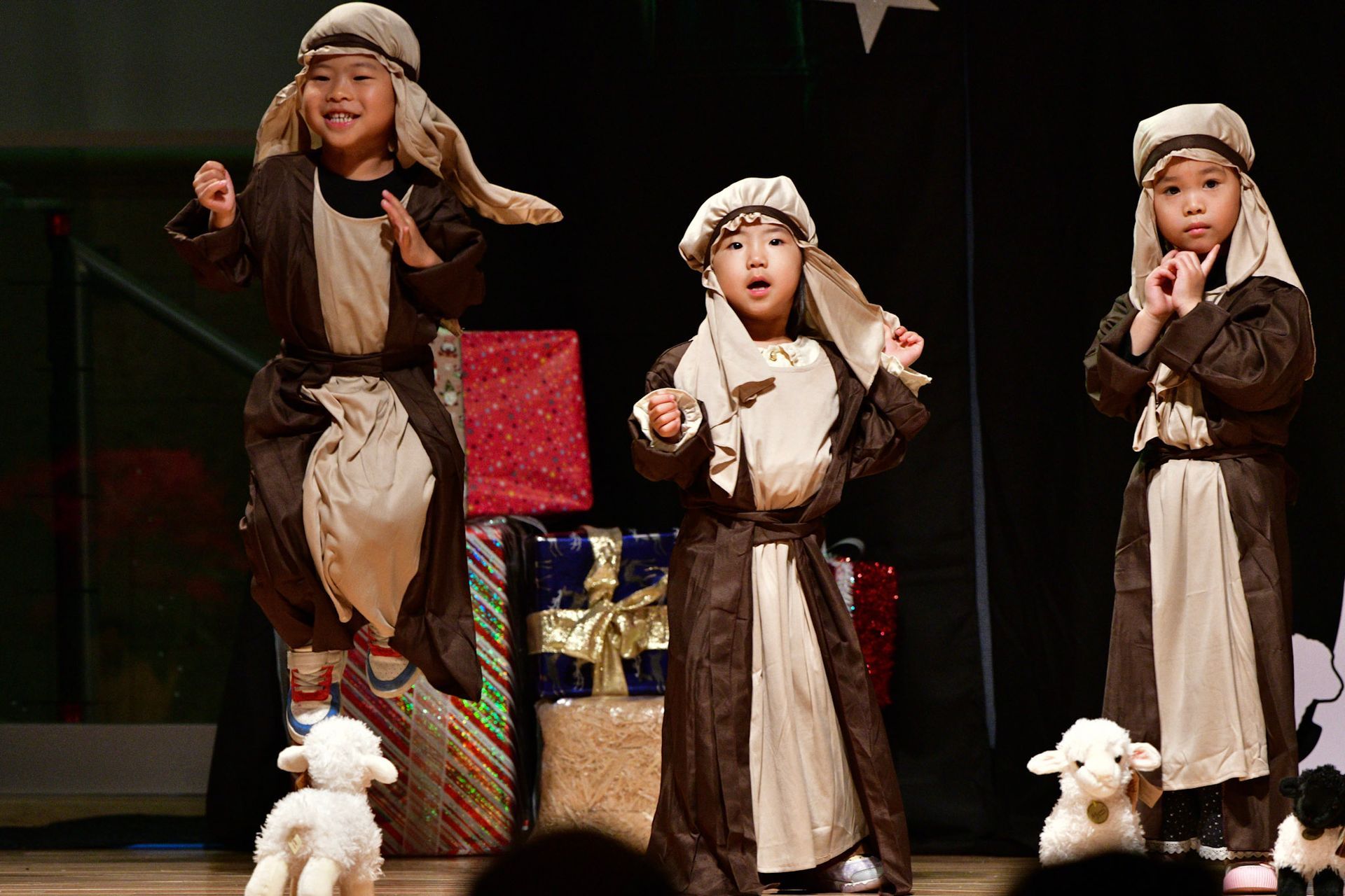 Three children in shepherd costumes on stage, singing near gifts and toy lambs.