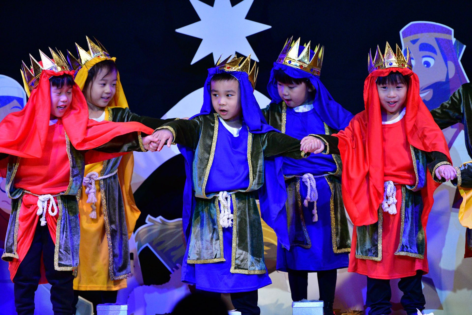 Children in costumes performing on a stage, with crowns and colored robes. Background has a star.