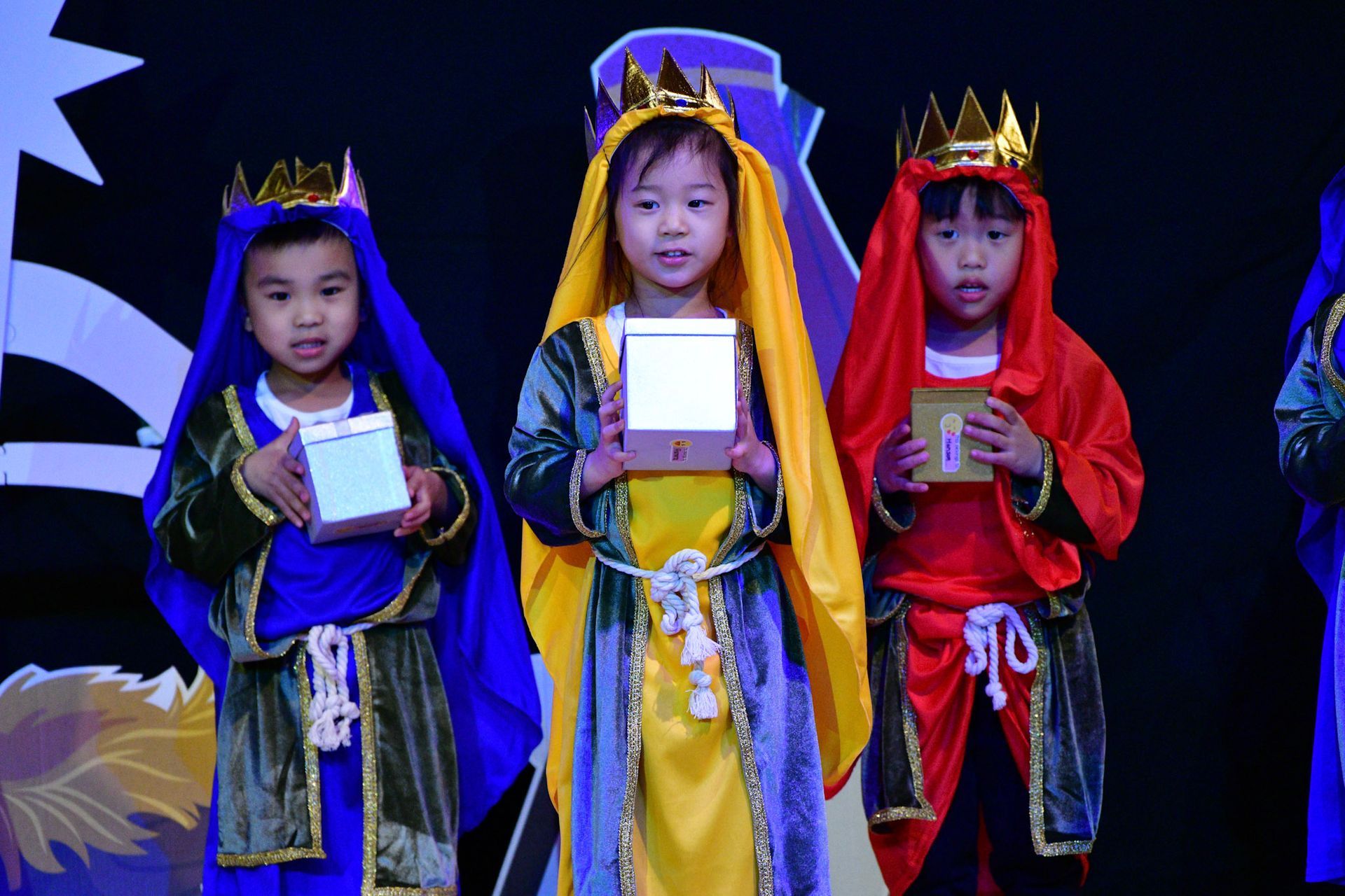 Three children in costumes, wearing crowns and holding gifts on stage.