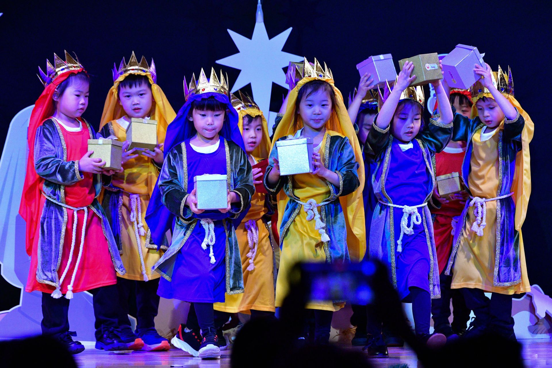 Children in costumes holding gifts on stage with a star backdrop.