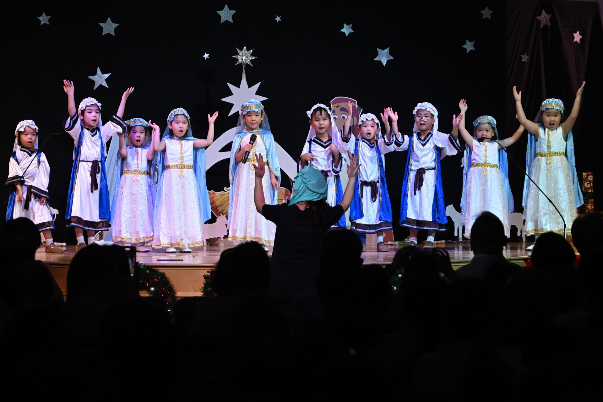 Children in costumes performing on stage, arms raised, star backdrop, audience visible.