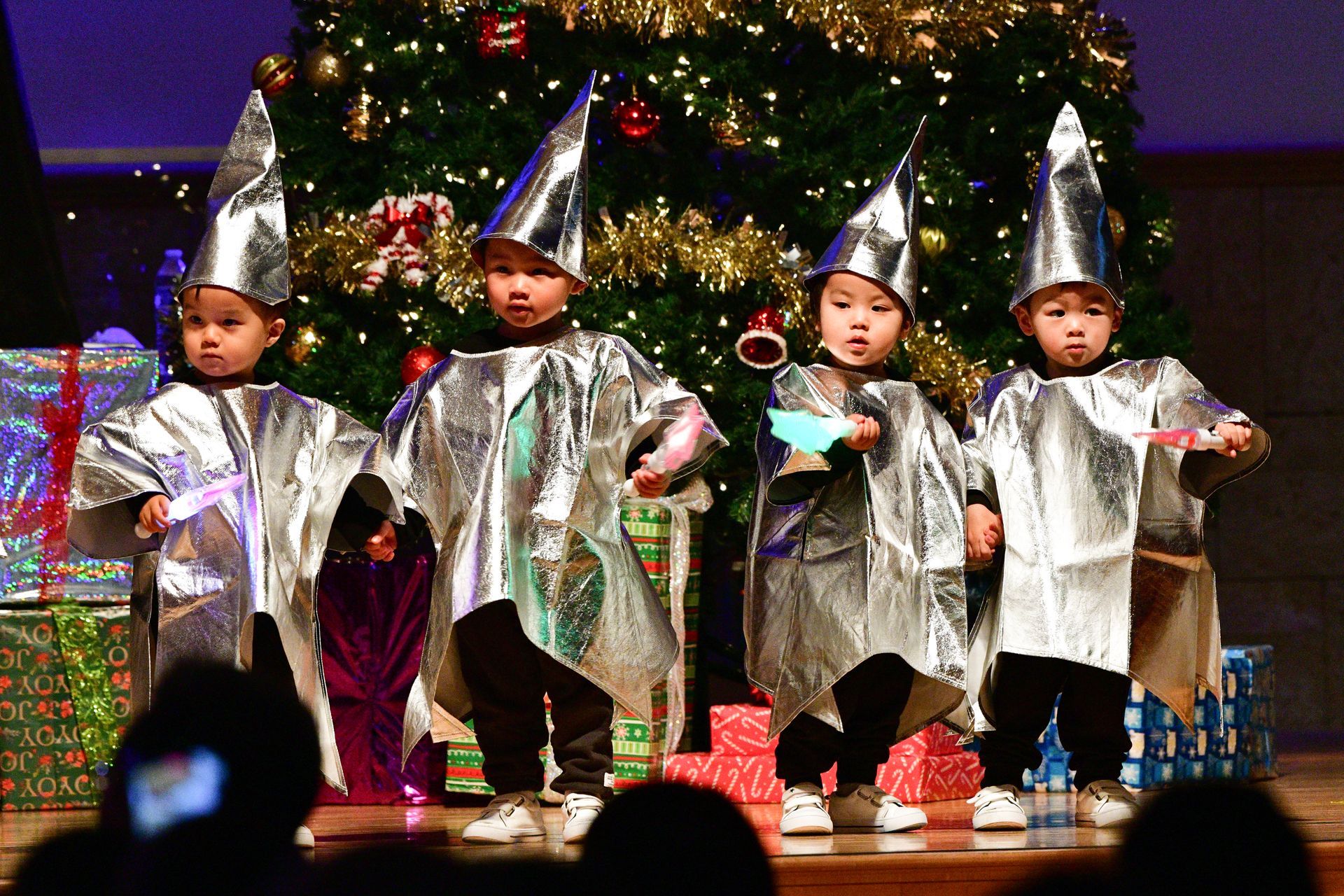 Children in silver costumes performing on stage in front of a Christmas tree.