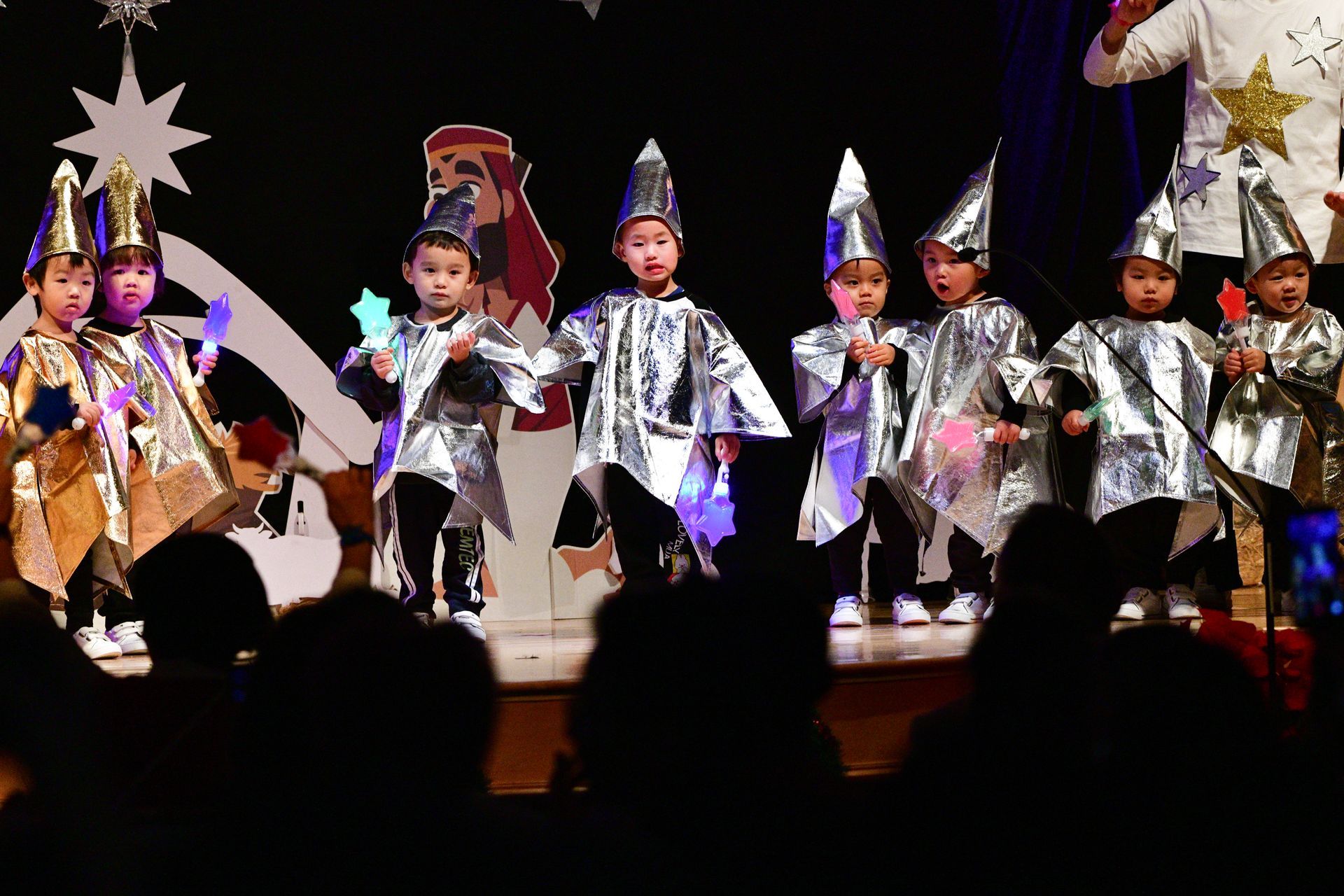 Children in silver costumes perform on stage, holding star-shaped props.