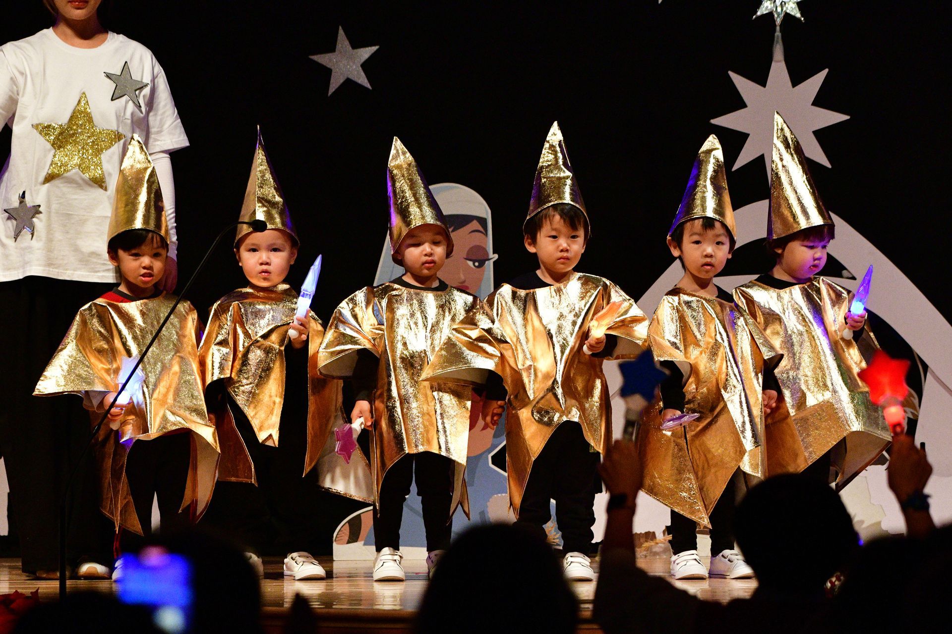 Children in gold wizard costumes on a stage with stars, holding wands, and performing.