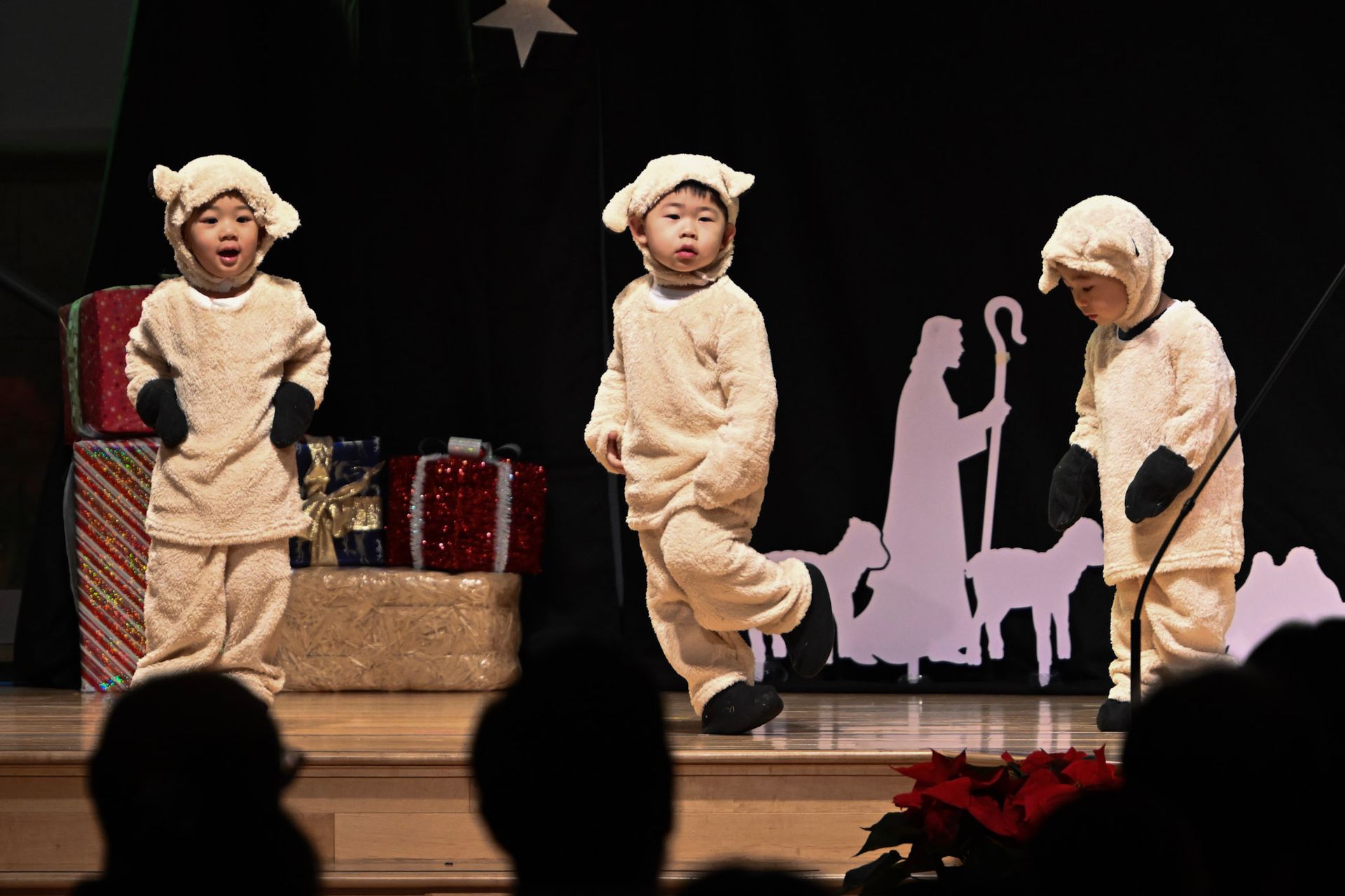 Three children in sheep costumes on stage. They are performing in front of a nativity scene cutout and present props.