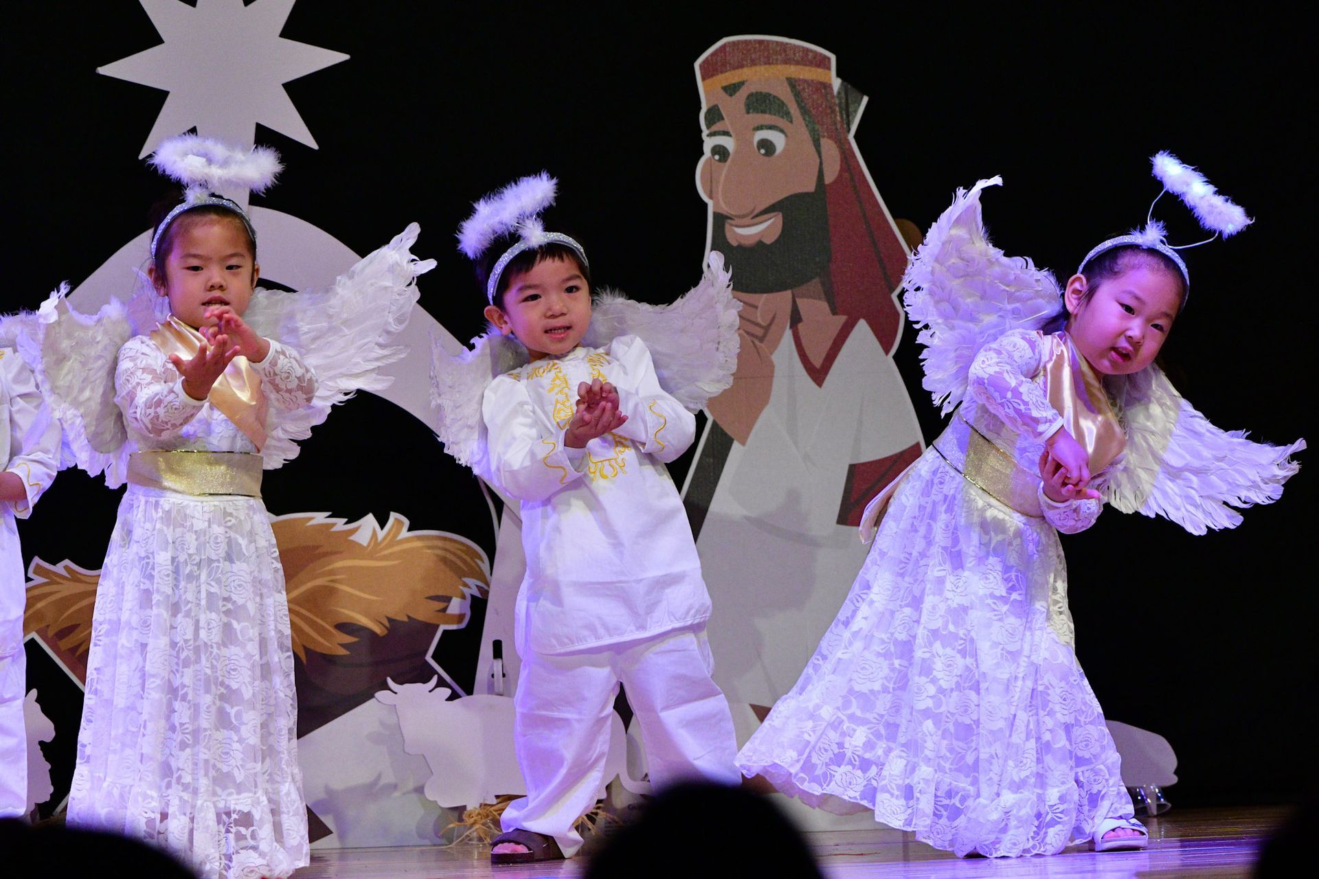 Children in angel costumes perform on stage, background with star and cartoon figure.