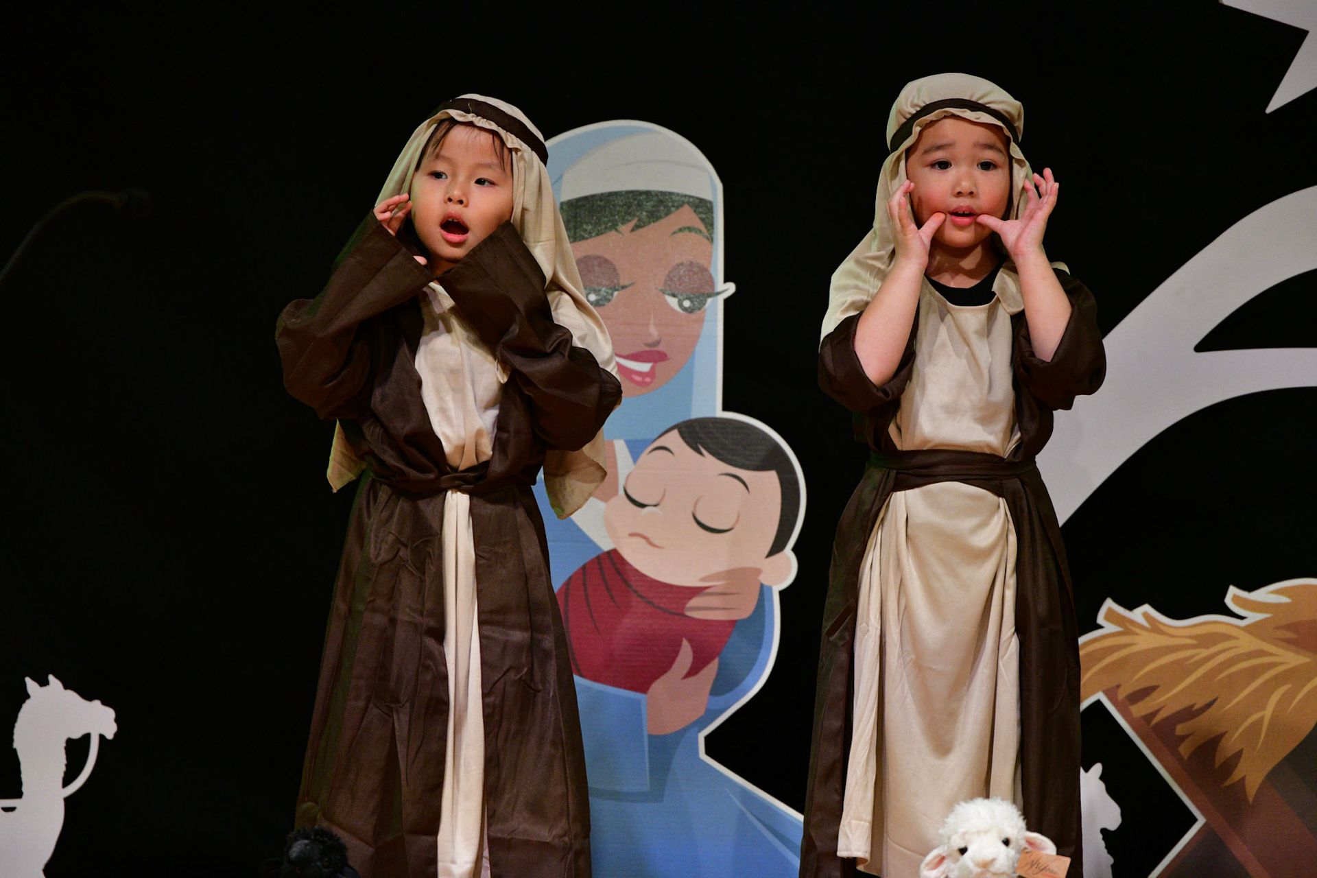 Two children in costumes perform a nativity play, with a backdrop of Mary and baby Jesus.