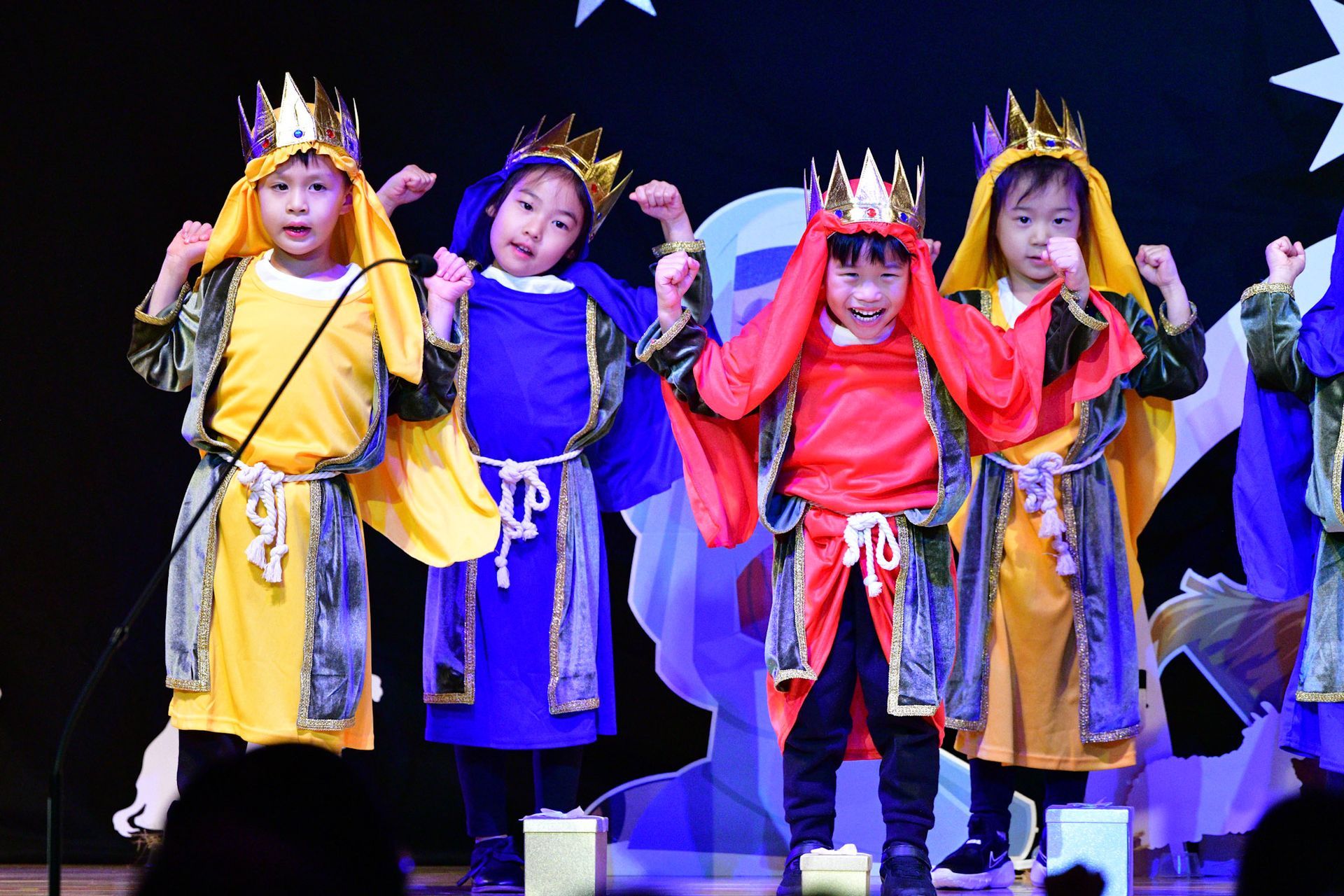 Children in colorful robes and crowns perform on stage, gold boxes present.