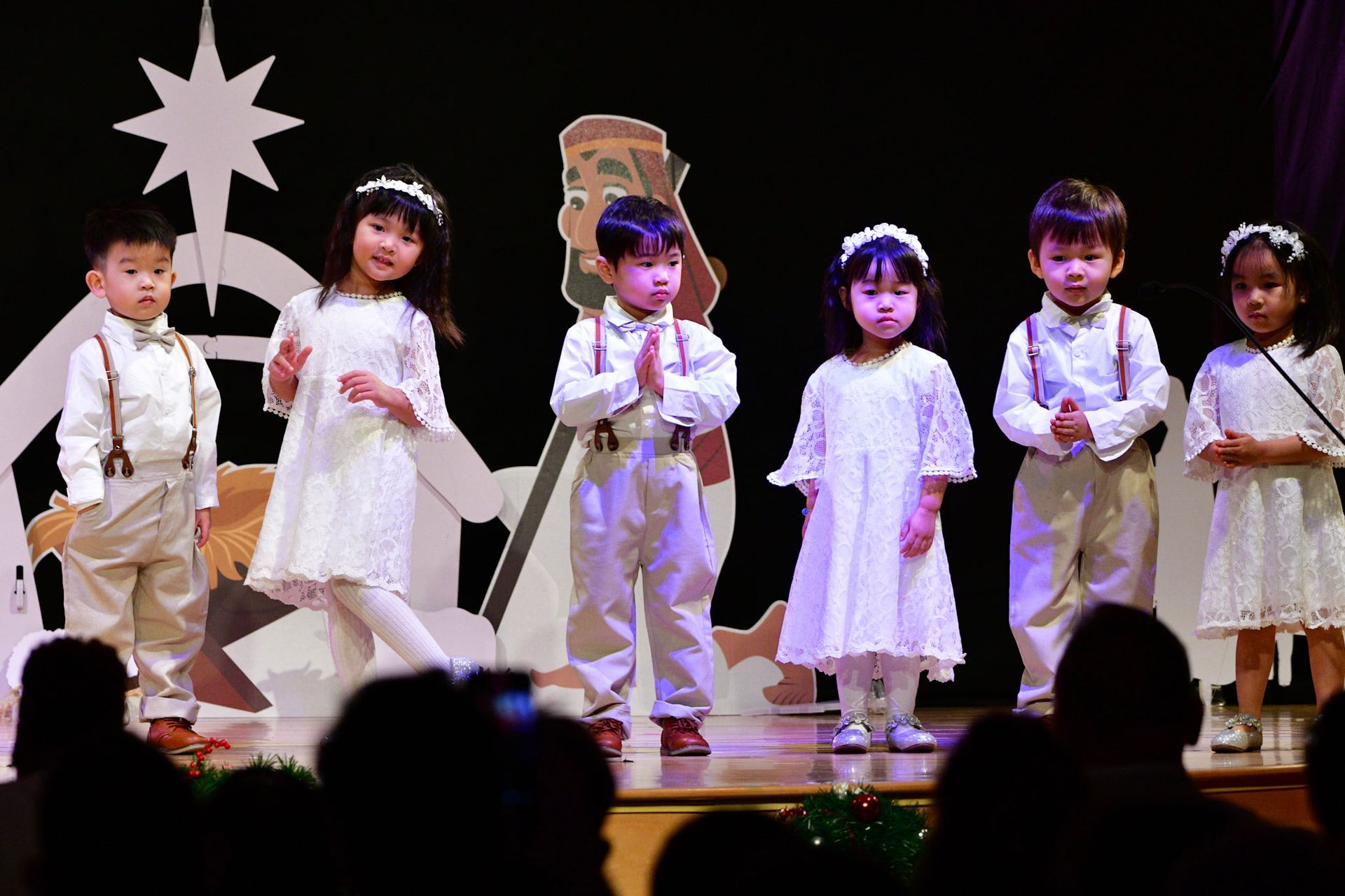 Children performing on stage, wearing white outfits and headbands. Christmas star in backdrop.