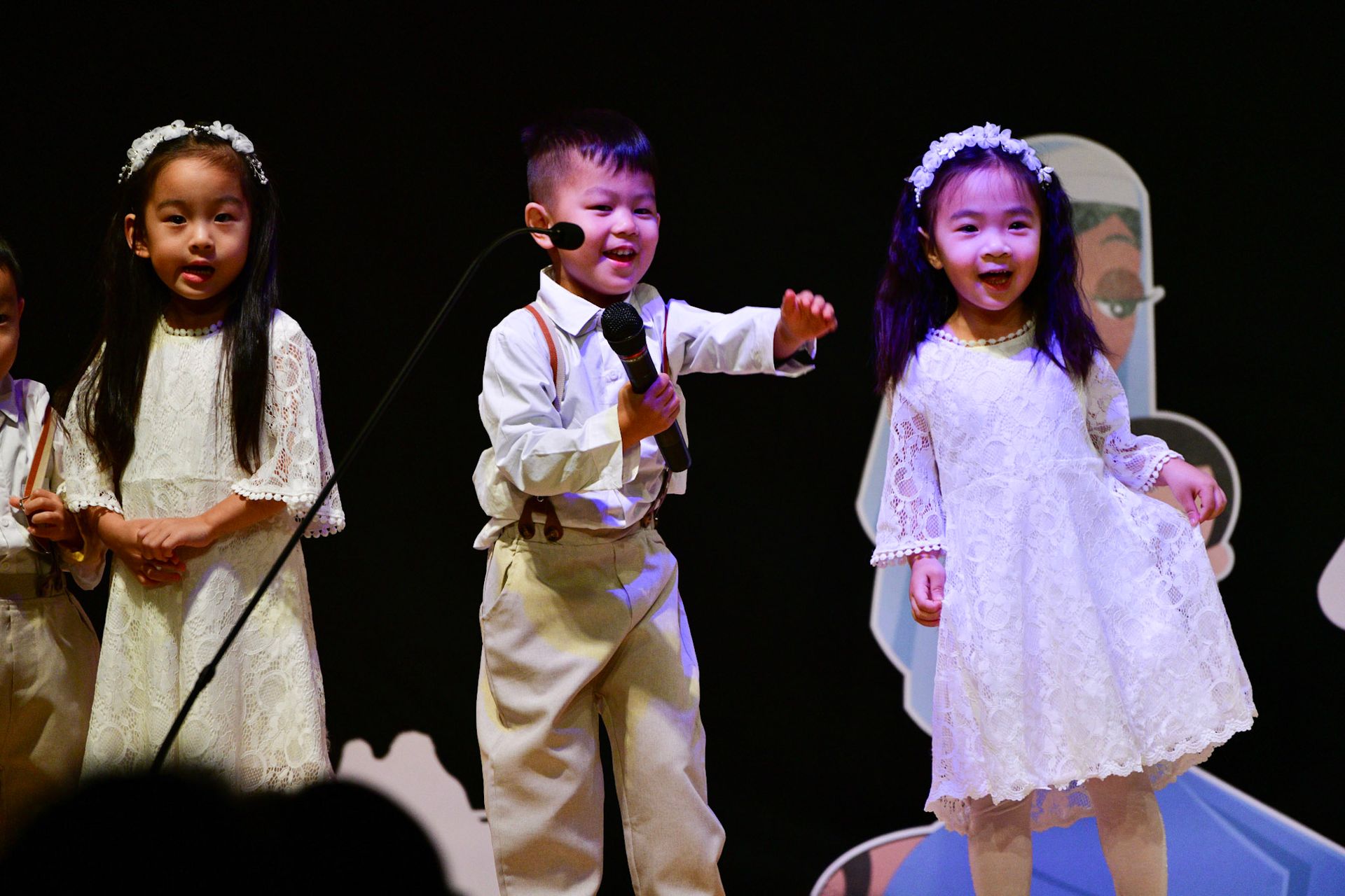 Children performing on stage, wearing white dresses and suits. One child holds a microphone and gestures.