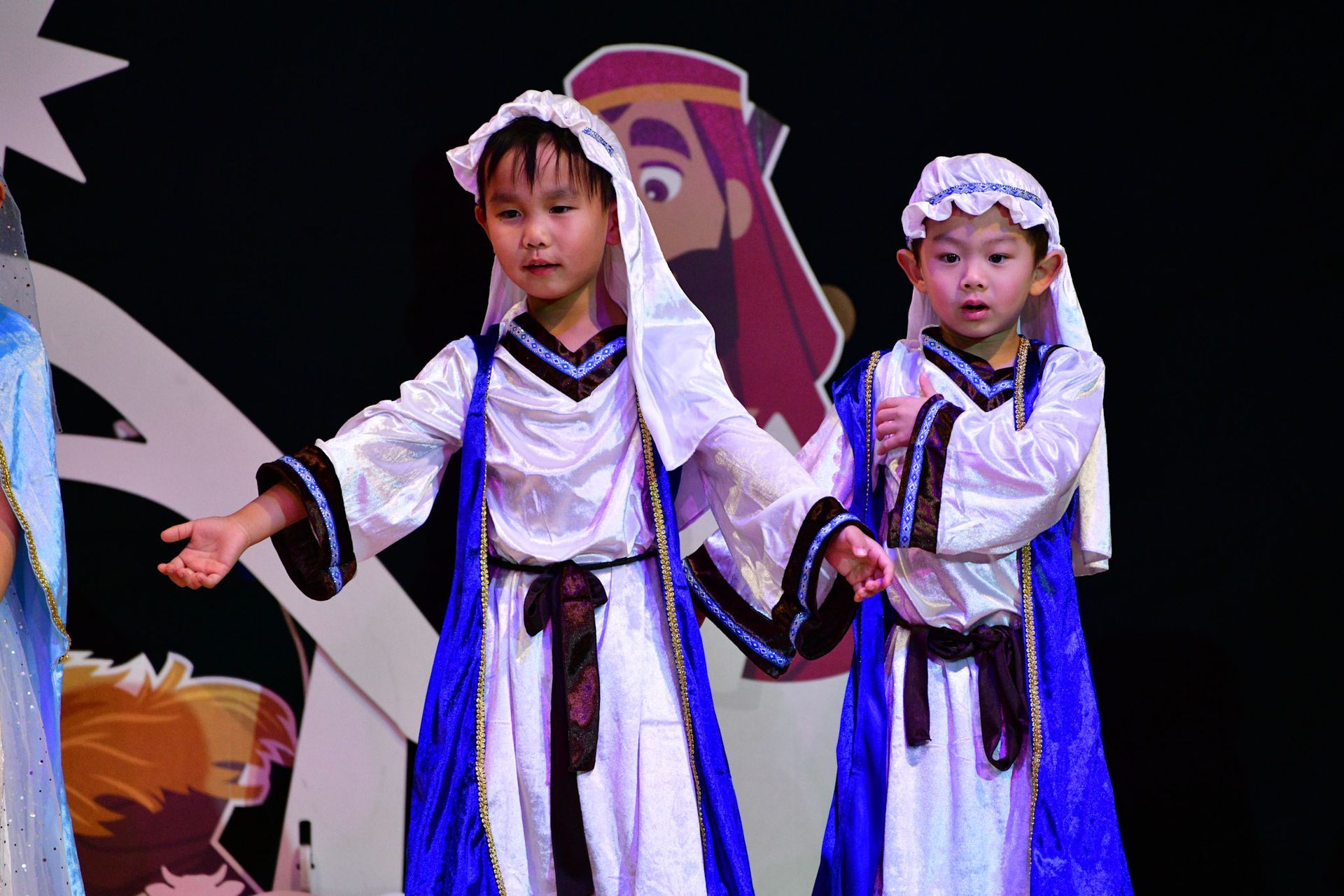 Children in blue and white robes perform on stage, with a cartoon background.