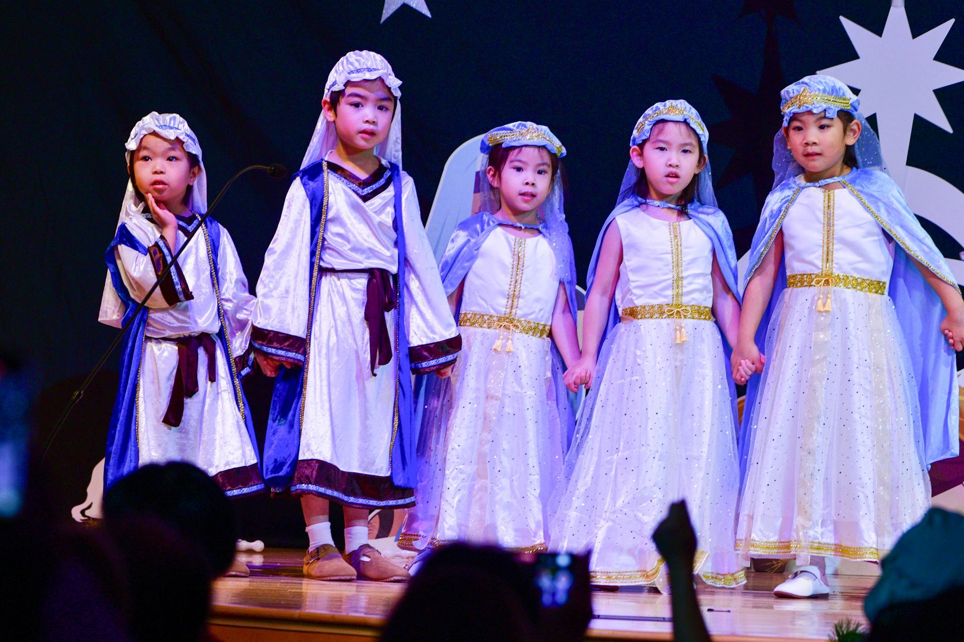 Children in costumes, holding hands on a stage, possibly in a nativity play with starry backdrop.