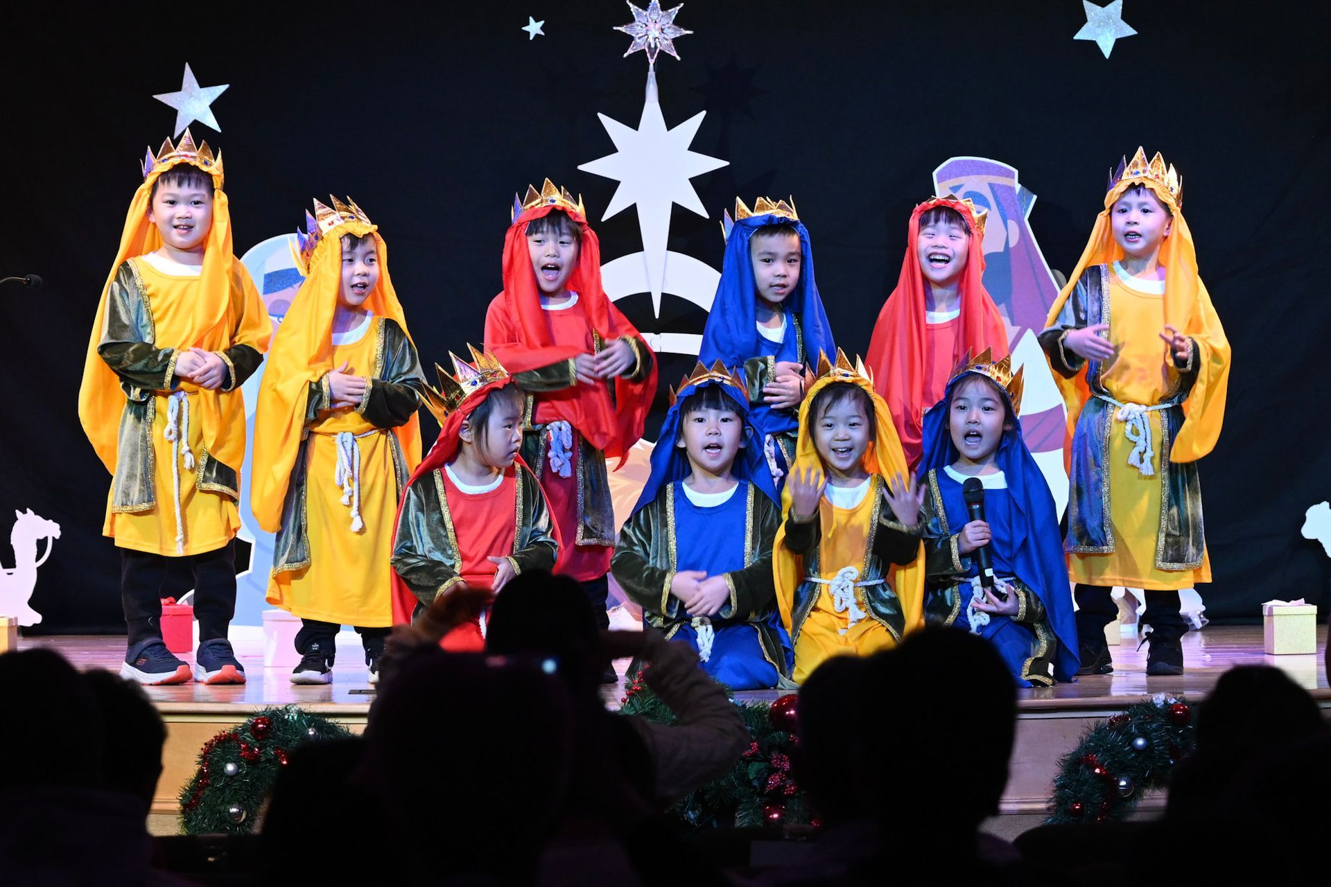 Children in colorful costumes sing on a stage with a starry backdrop.