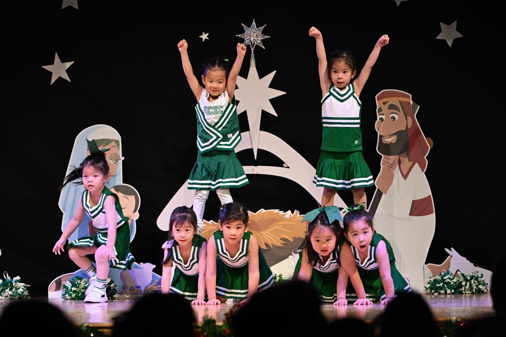 Children in green and white cheerleading outfits performing on stage with religious-themed props.