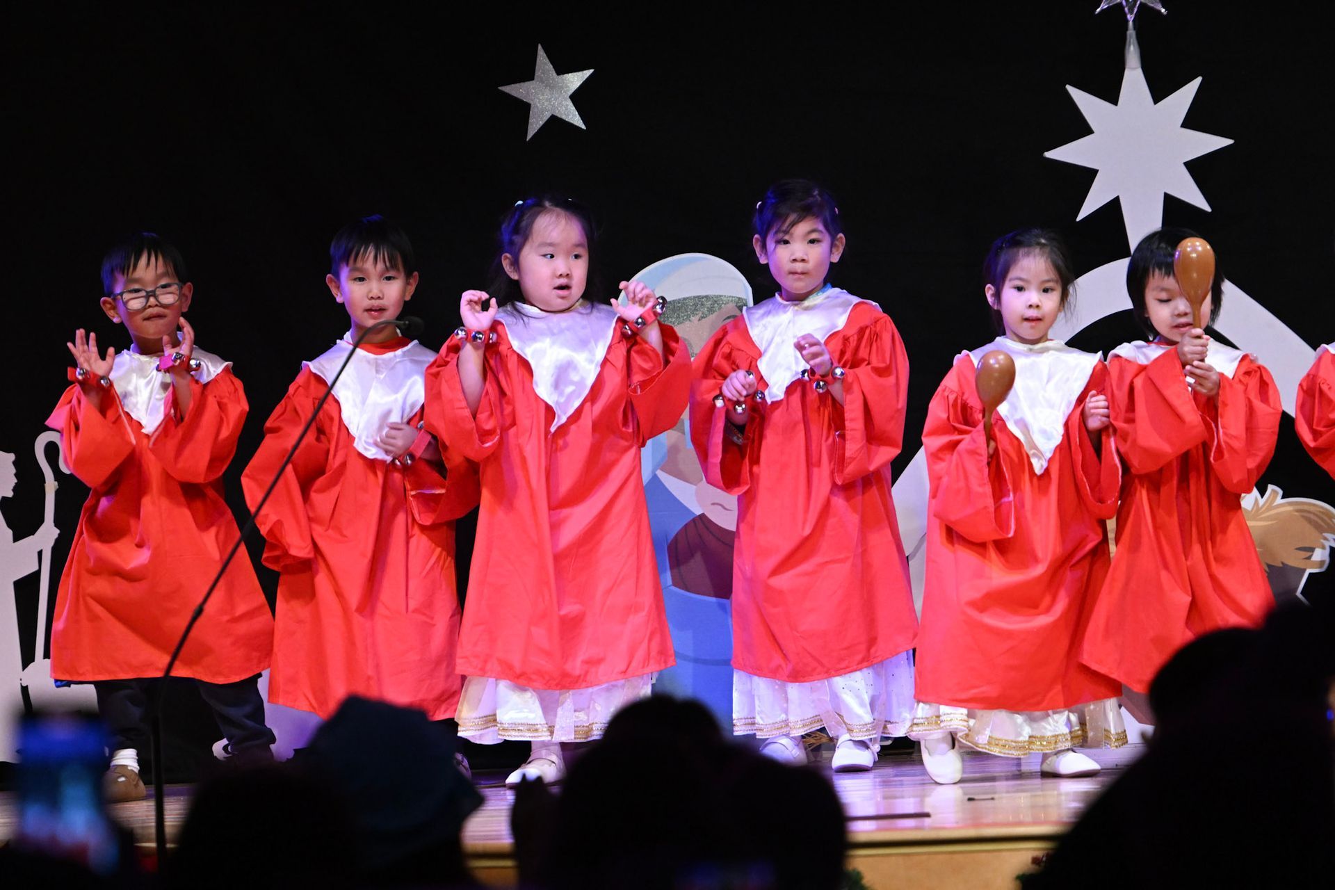 Children in red robes perform on stage, Christmas stars in background.