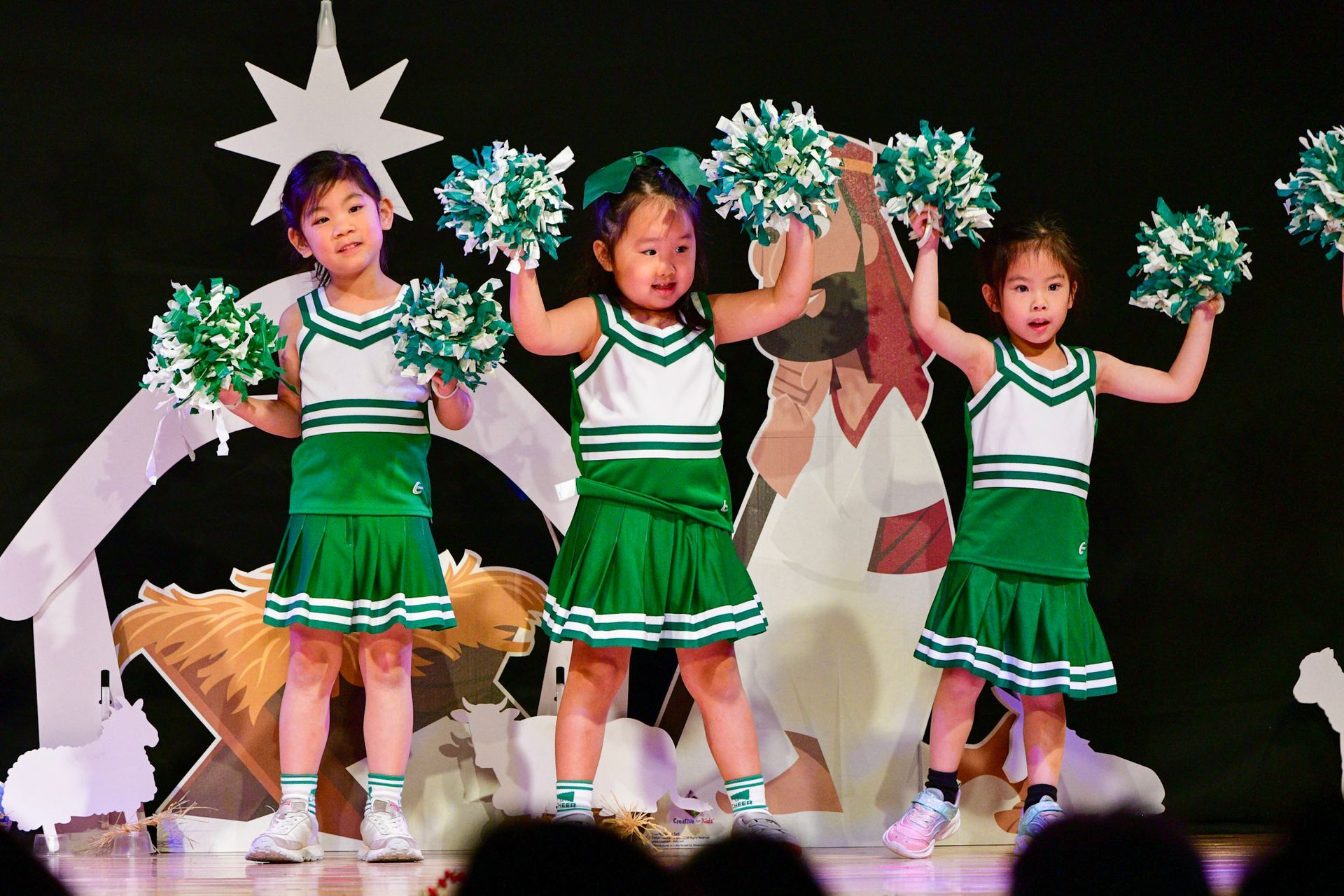 Three young girls in green and white cheerleading outfits with pom-poms perform on stage.