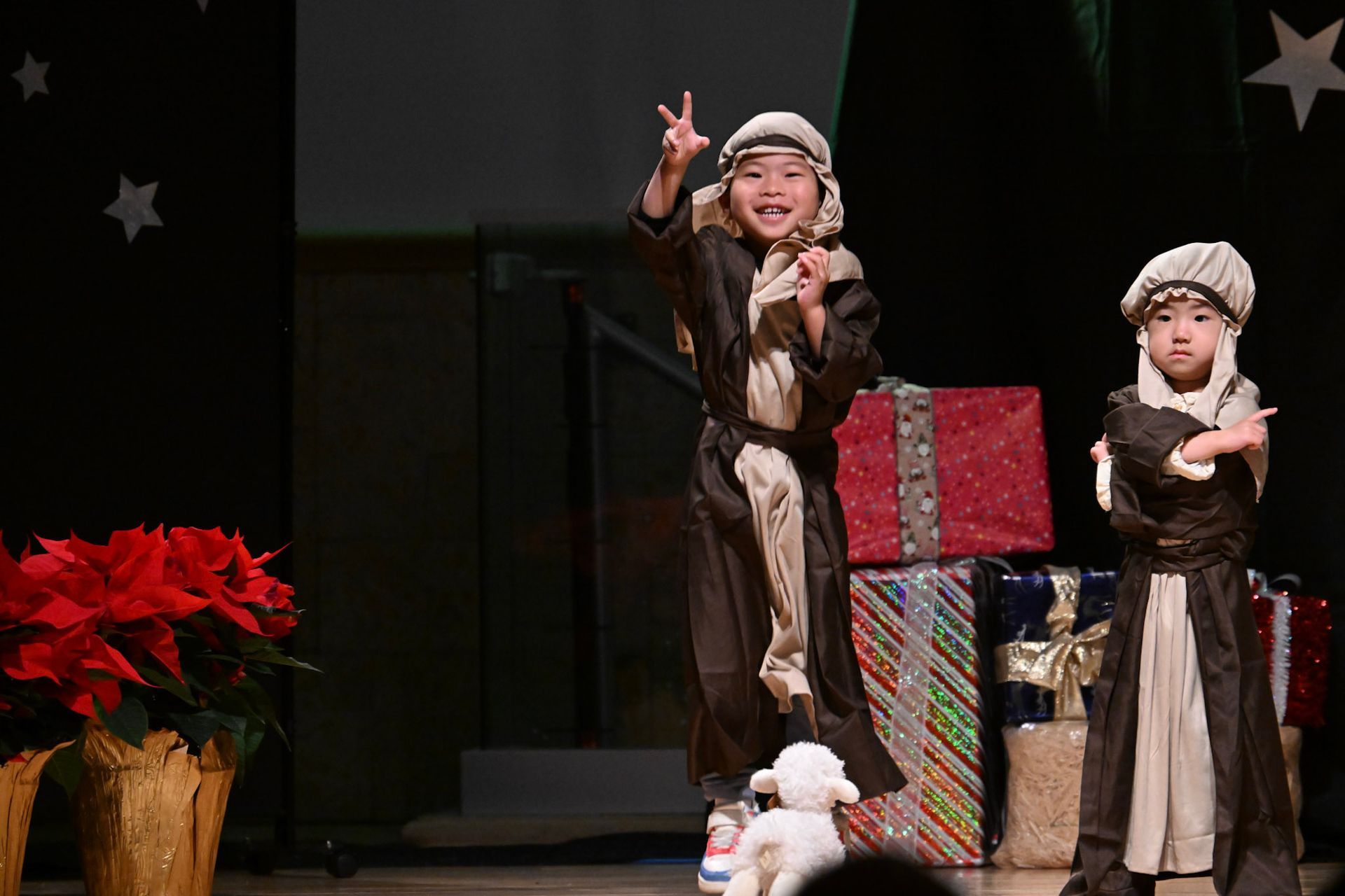 Two children dressed as shepherds on stage with presents and a lamb. One makes a peace sign.