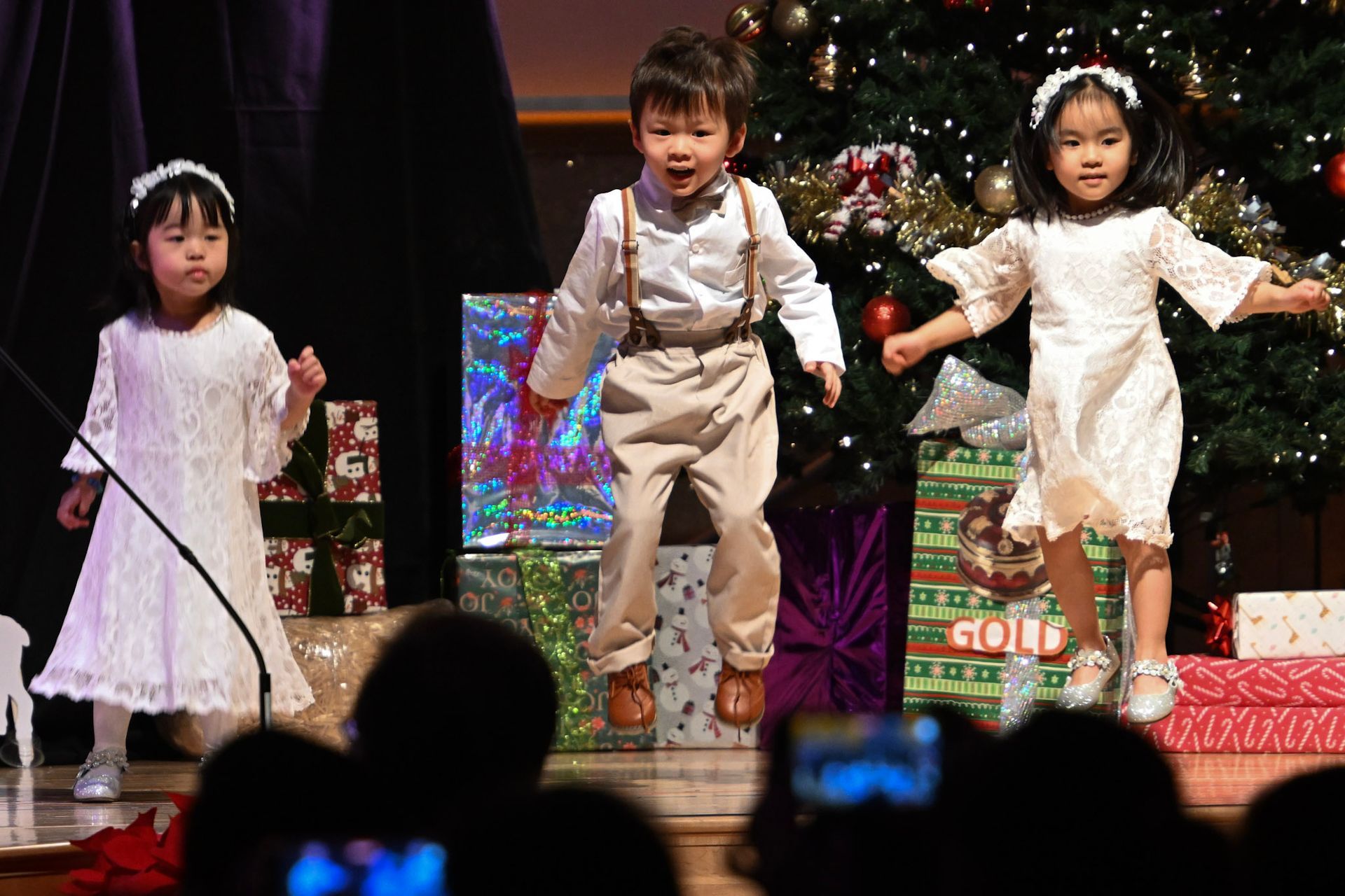Three children performing on stage in front of a Christmas tree and wrapped presents.