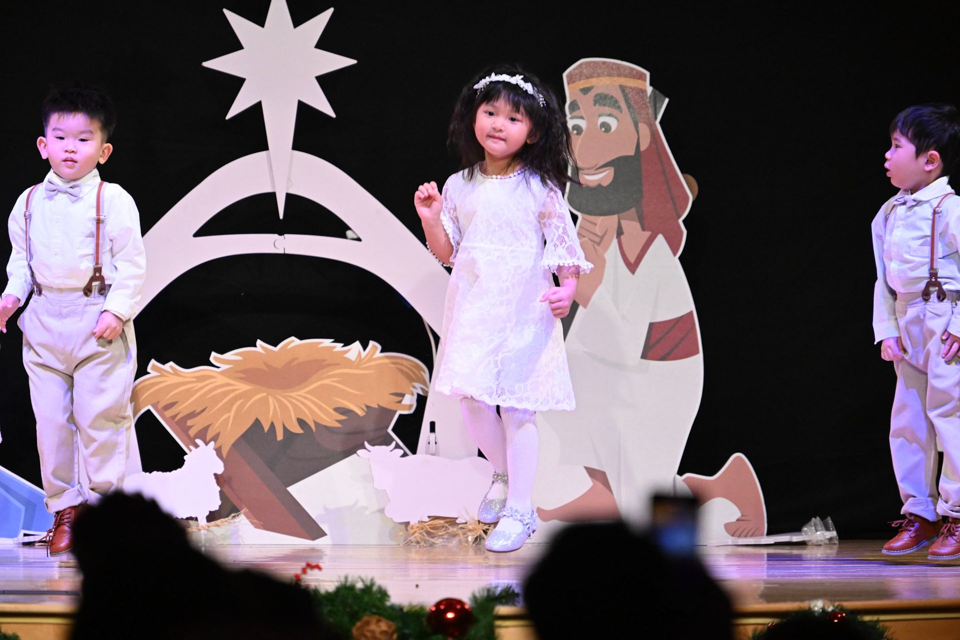 Children performing a nativity scene on stage, with paper cutouts of a manger and a person.