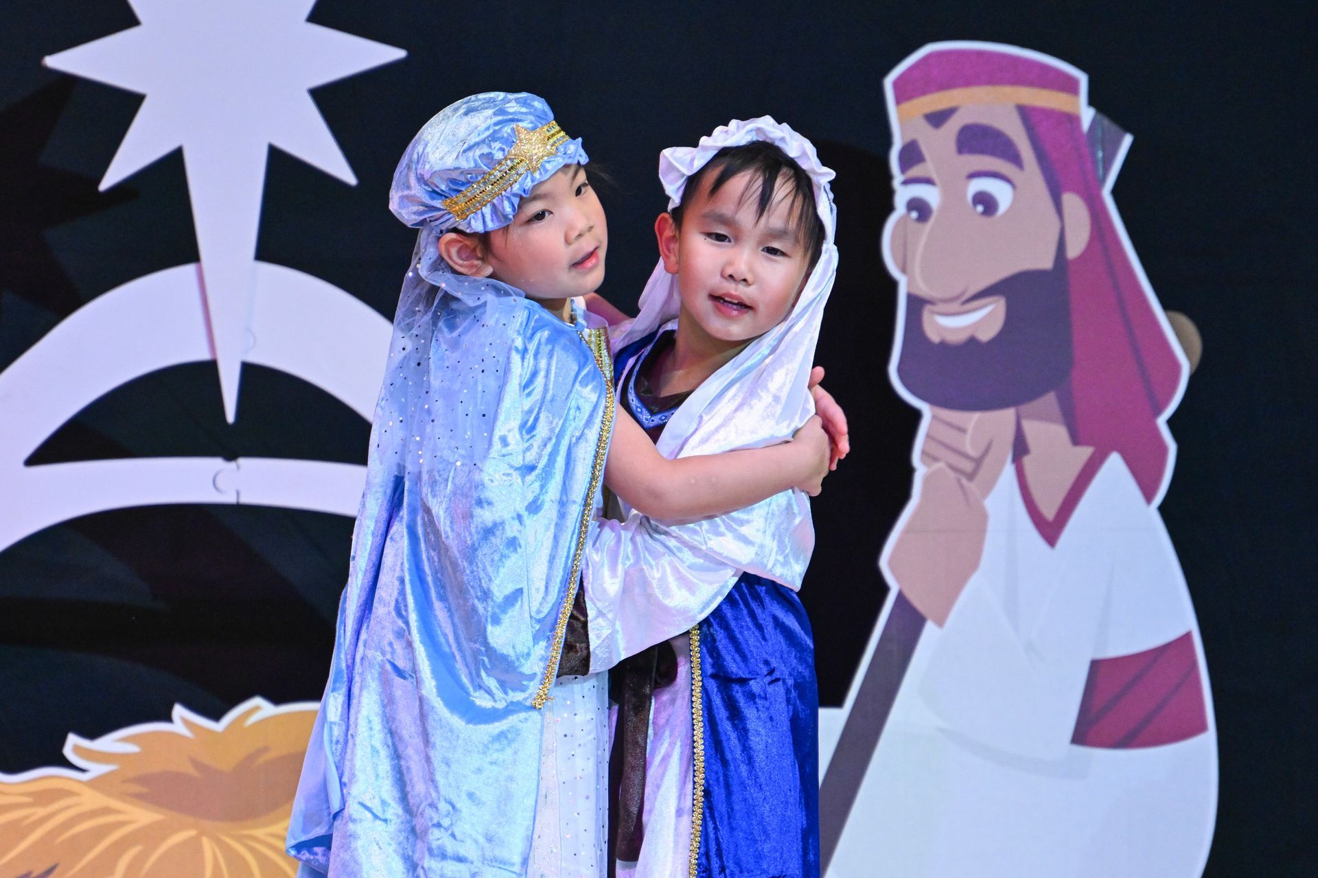Children in costumes hugging, with a cartoon cutout of a man. Christmas-themed stage setting, star in the background.