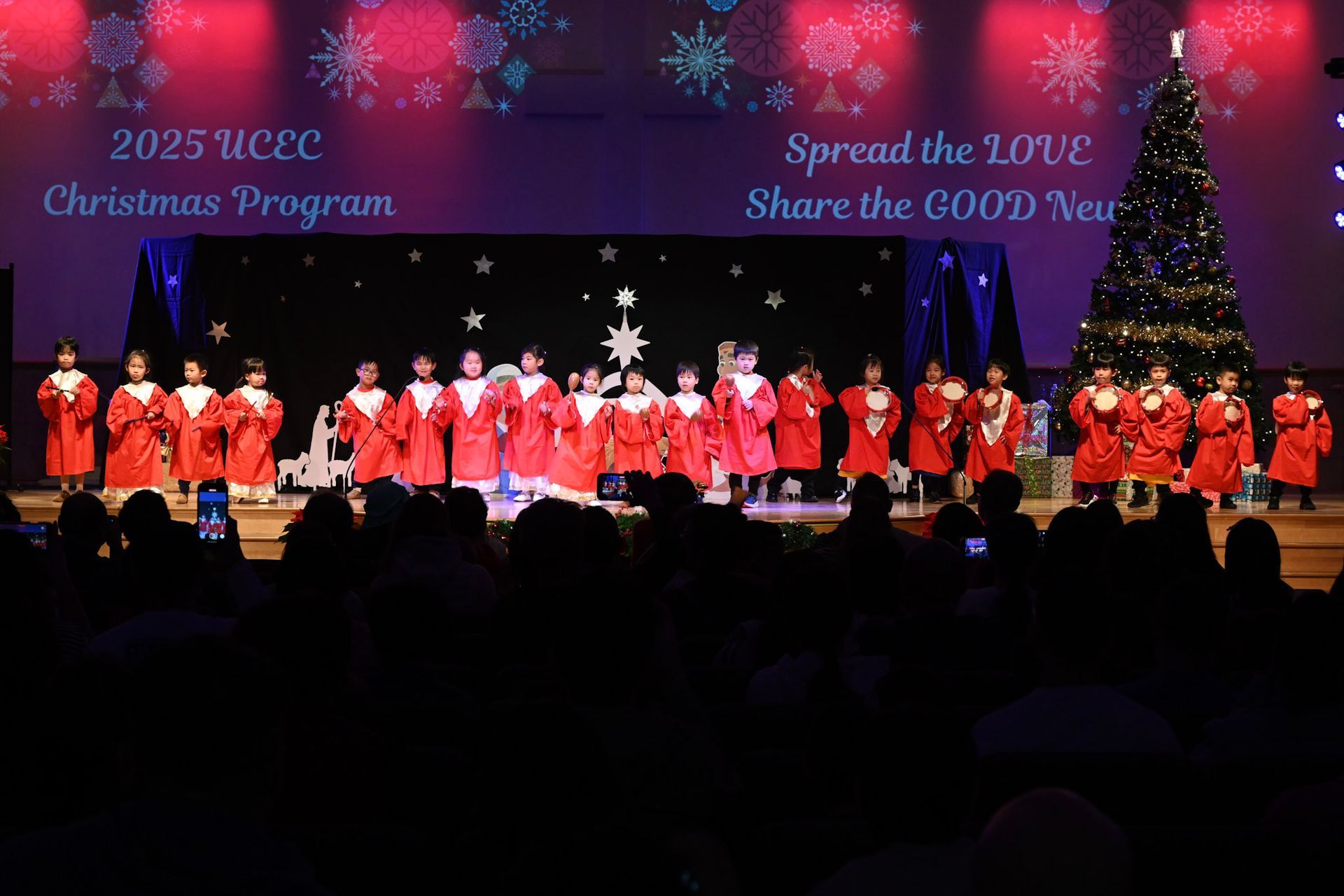 Children in red robes on stage for a Christmas program, in front of a Christmas tree.