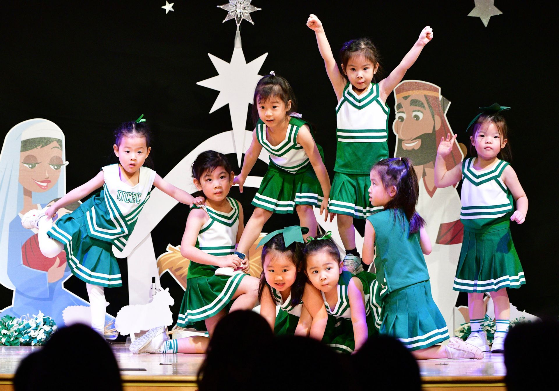 Children in green and white cheerleading outfits perform on stage with Mary and Jesus cutouts.