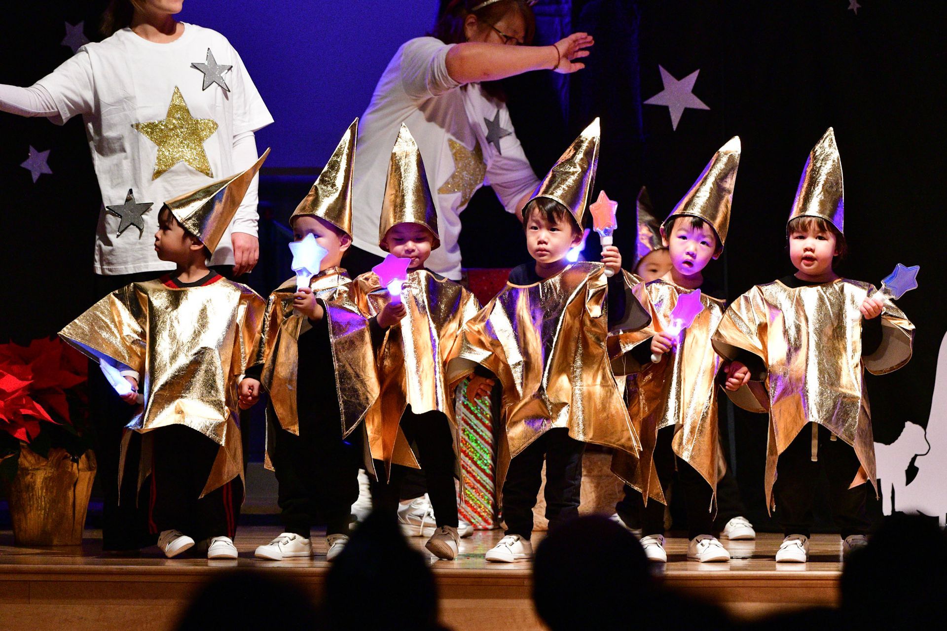 Children in gold costumes and hats perform on a stage with two adults.