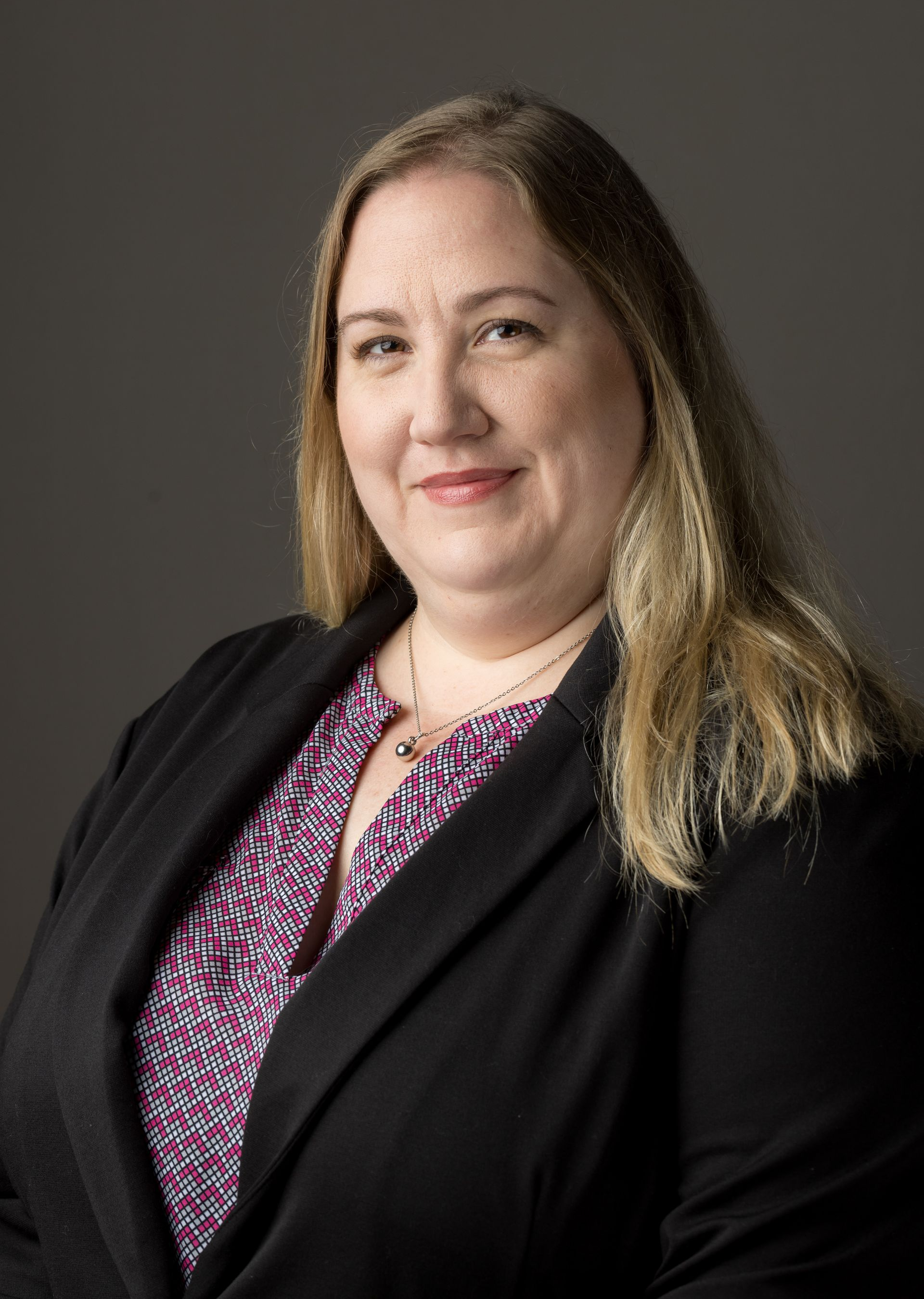 Woman with blonde hair wearing a black blazer and patterned shirt smiles, looking at the camera.