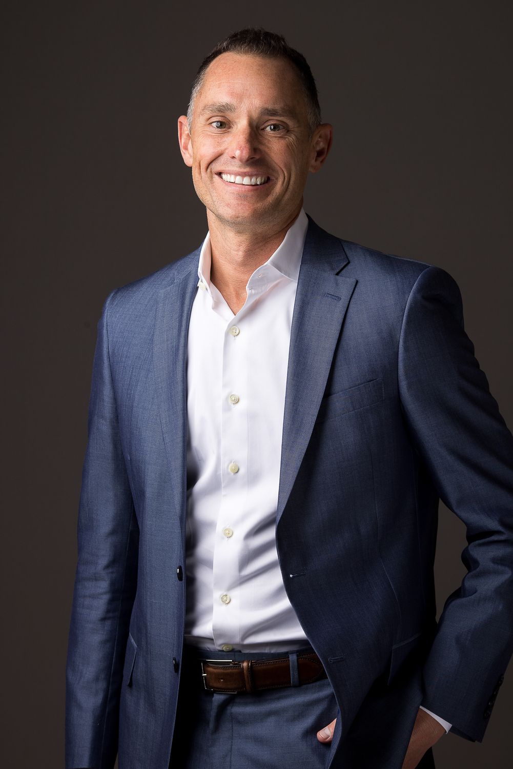 Man with short dark hair, smiling, wearing a blue blazer and white shirt against a gray background.