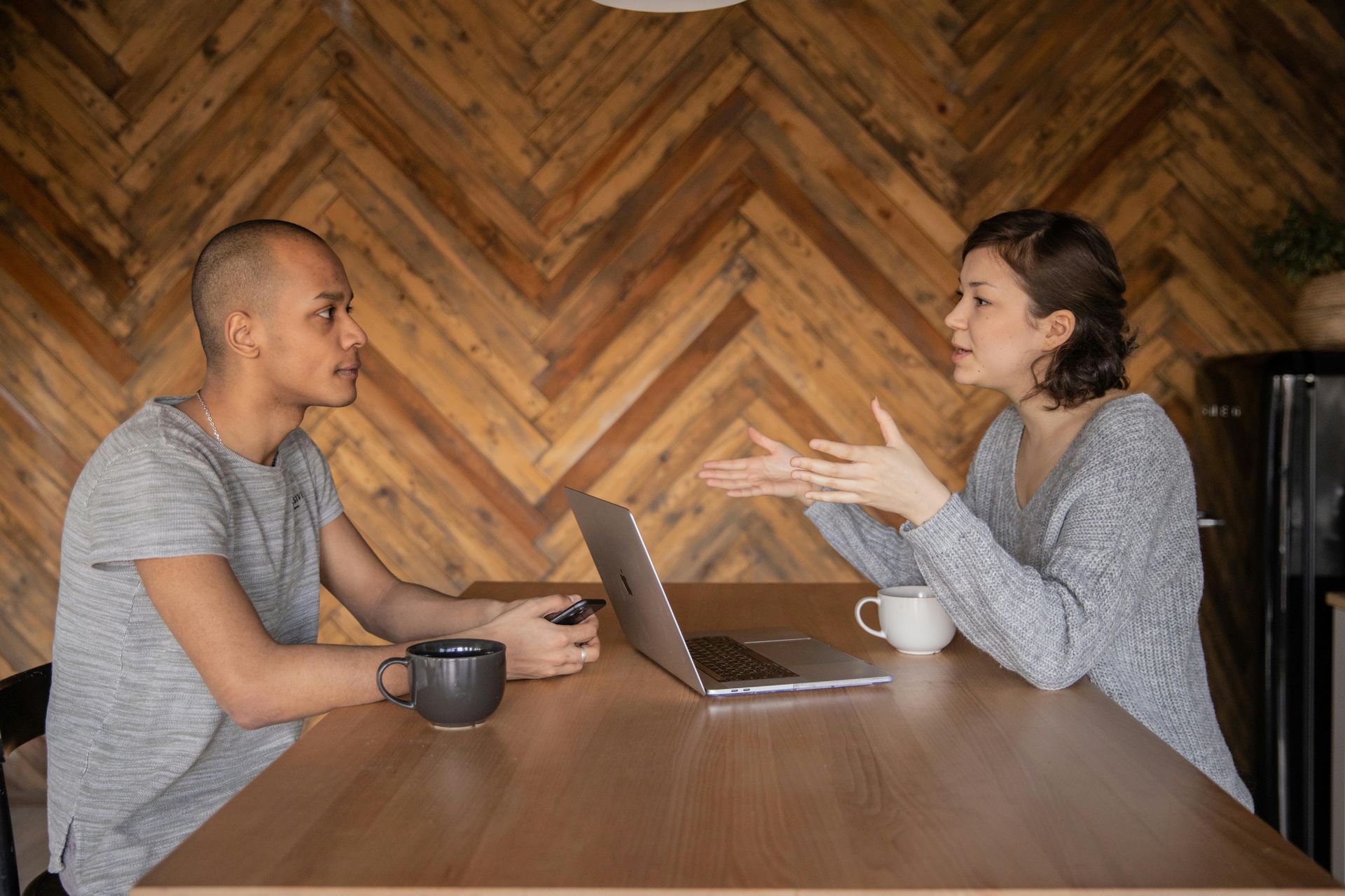 Man and woman sit at a table with a laptop, mugs. Woman gestures while speaking, man listens. Wooden background.
