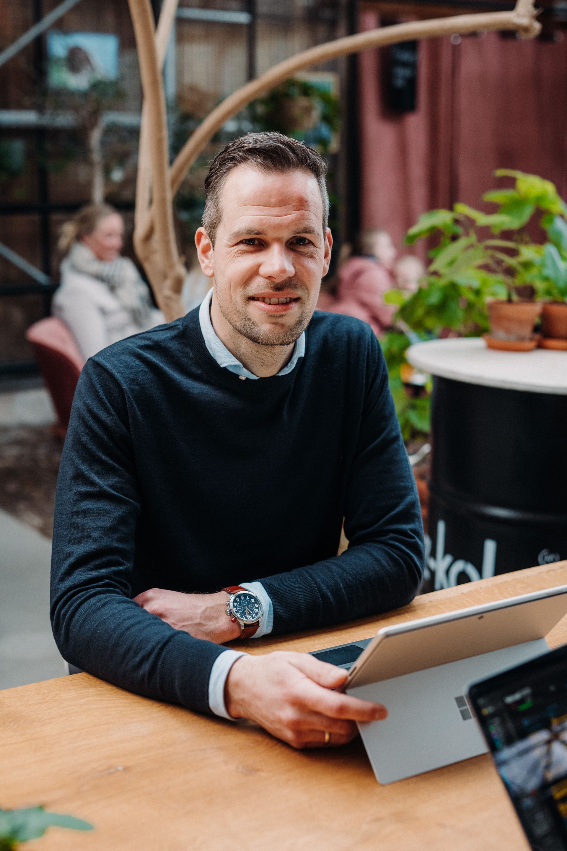 Een man in een donkerblauwe trui zit aan een tafel met een vrouw op de achtergrond.