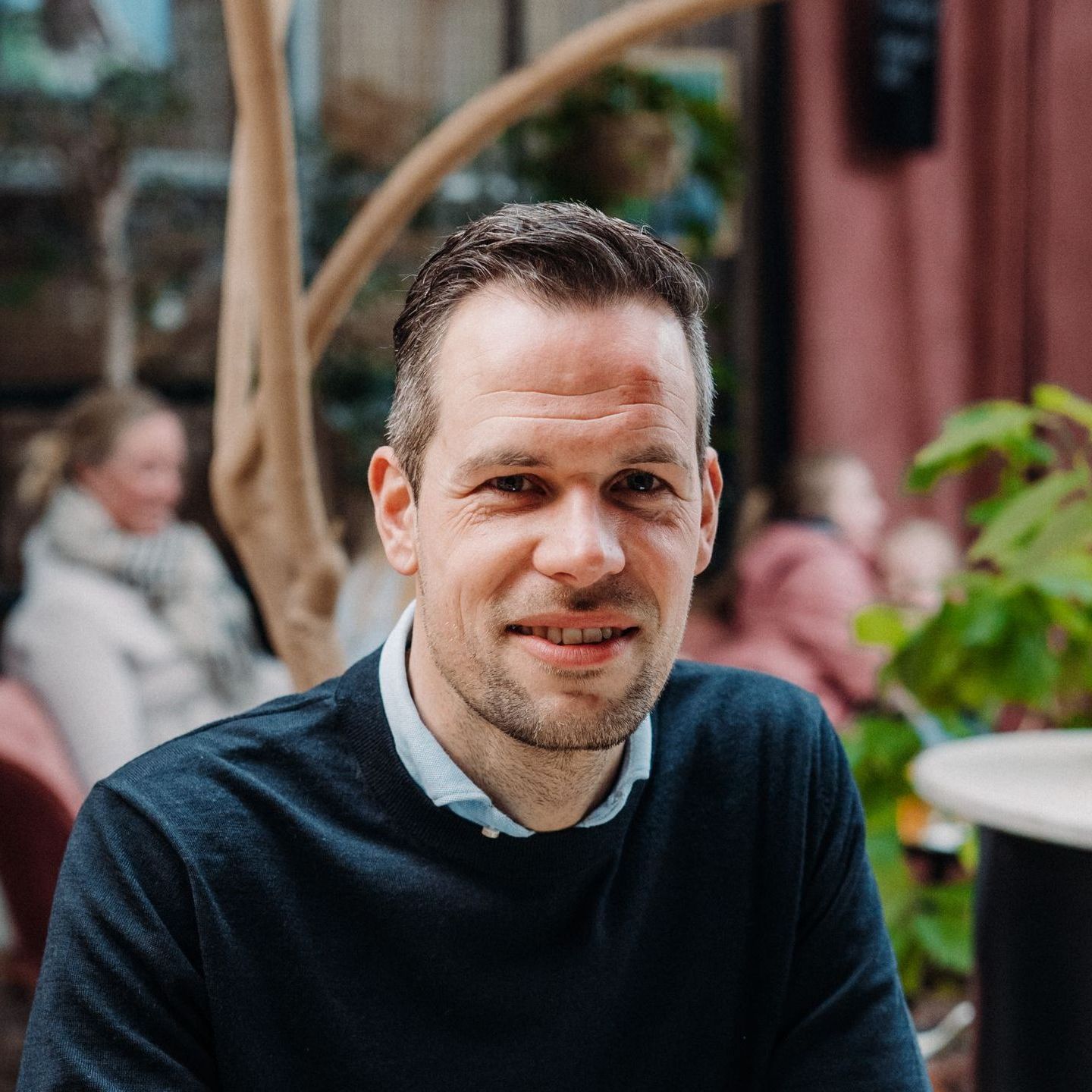 Een man in een donkerblauwe trui zit aan een tafel met een vrouw op de achtergrond.