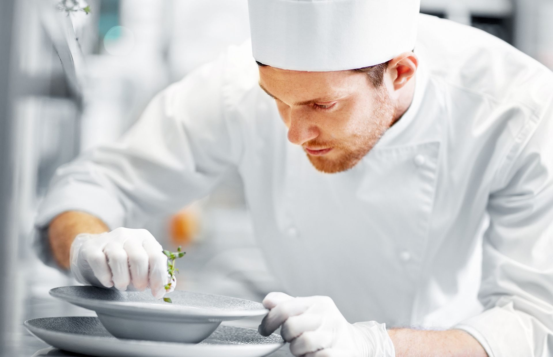 Chef meticulously garnishing a plate in a professional kitchen, wearing a white uniform and gloves.