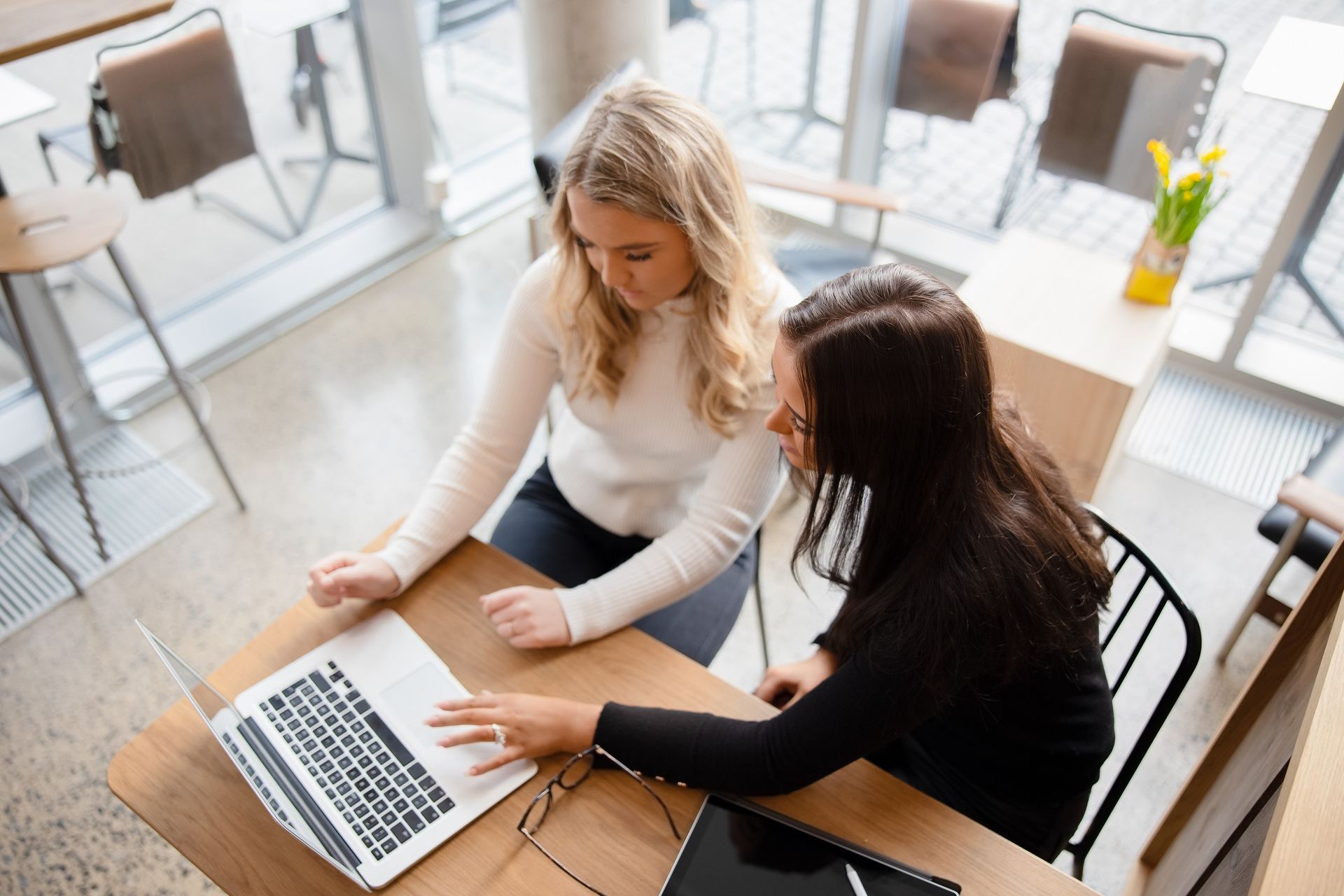 Two women looking at a laptop, seated at a wooden table in a cafe, one pointing at the screen.