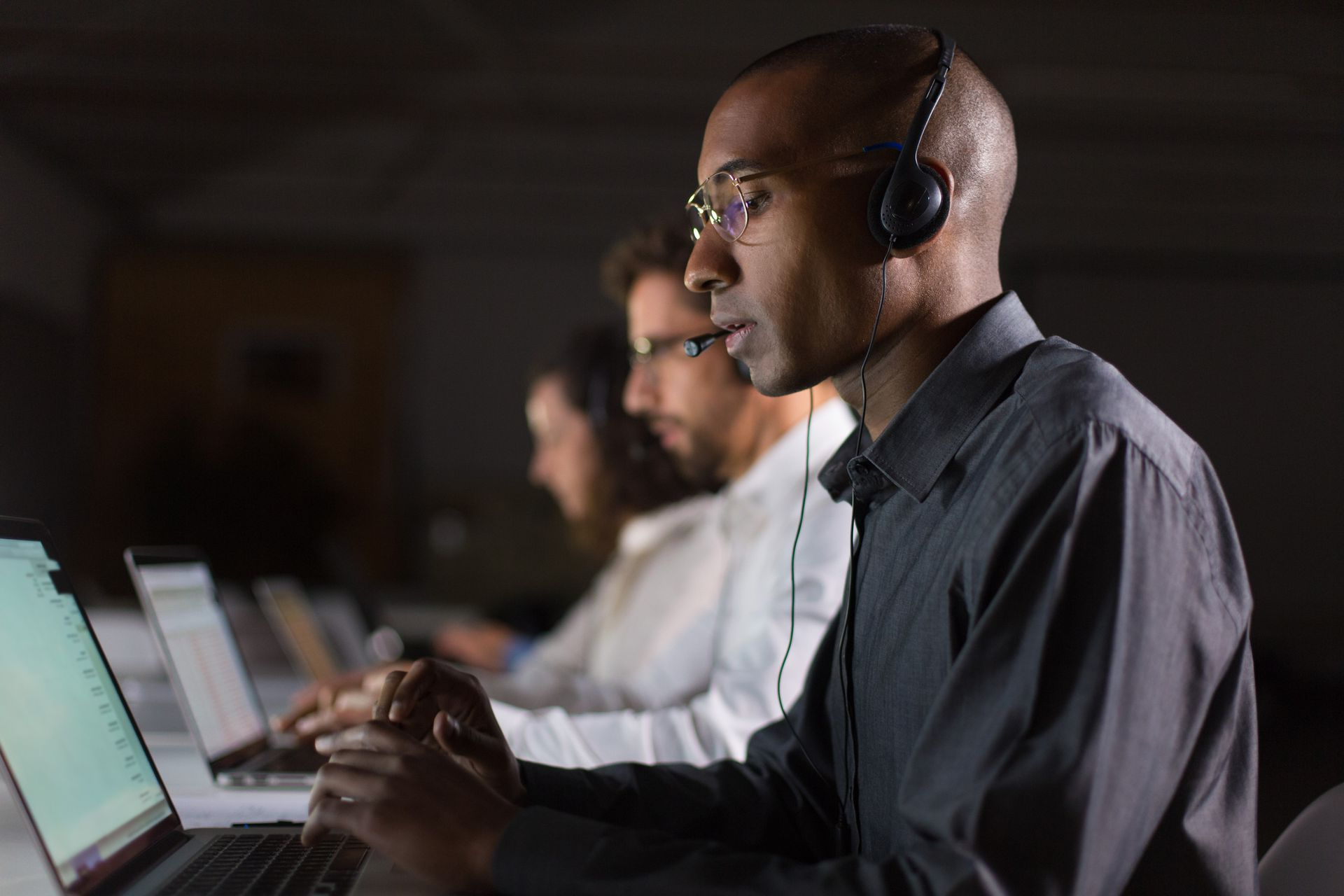 Man wearing headset and glasses, working on laptop in dimly lit office.