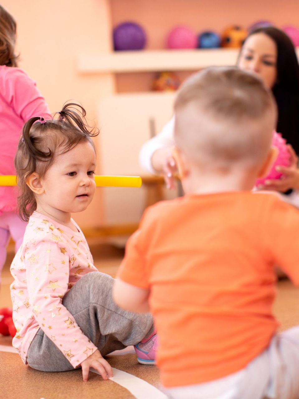 Children playing in a brightly lit room with a teacher, one girl stares towards the right.
