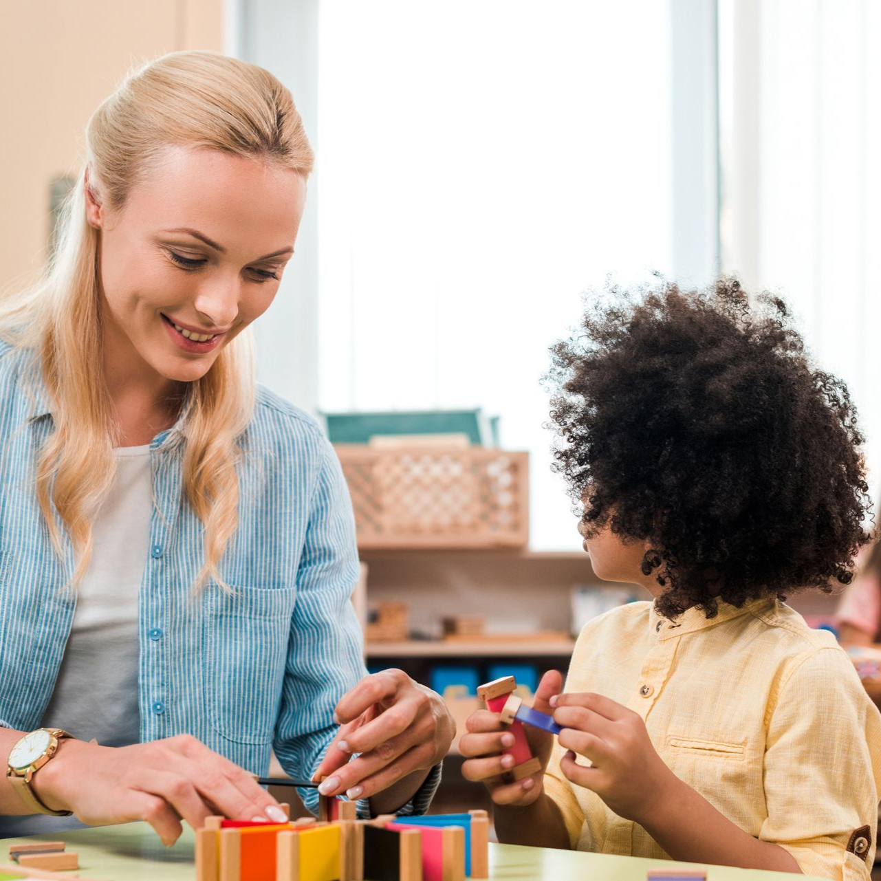 Teacher and child playing with colorful blocks in a classroom. The teacher smiles at the child.