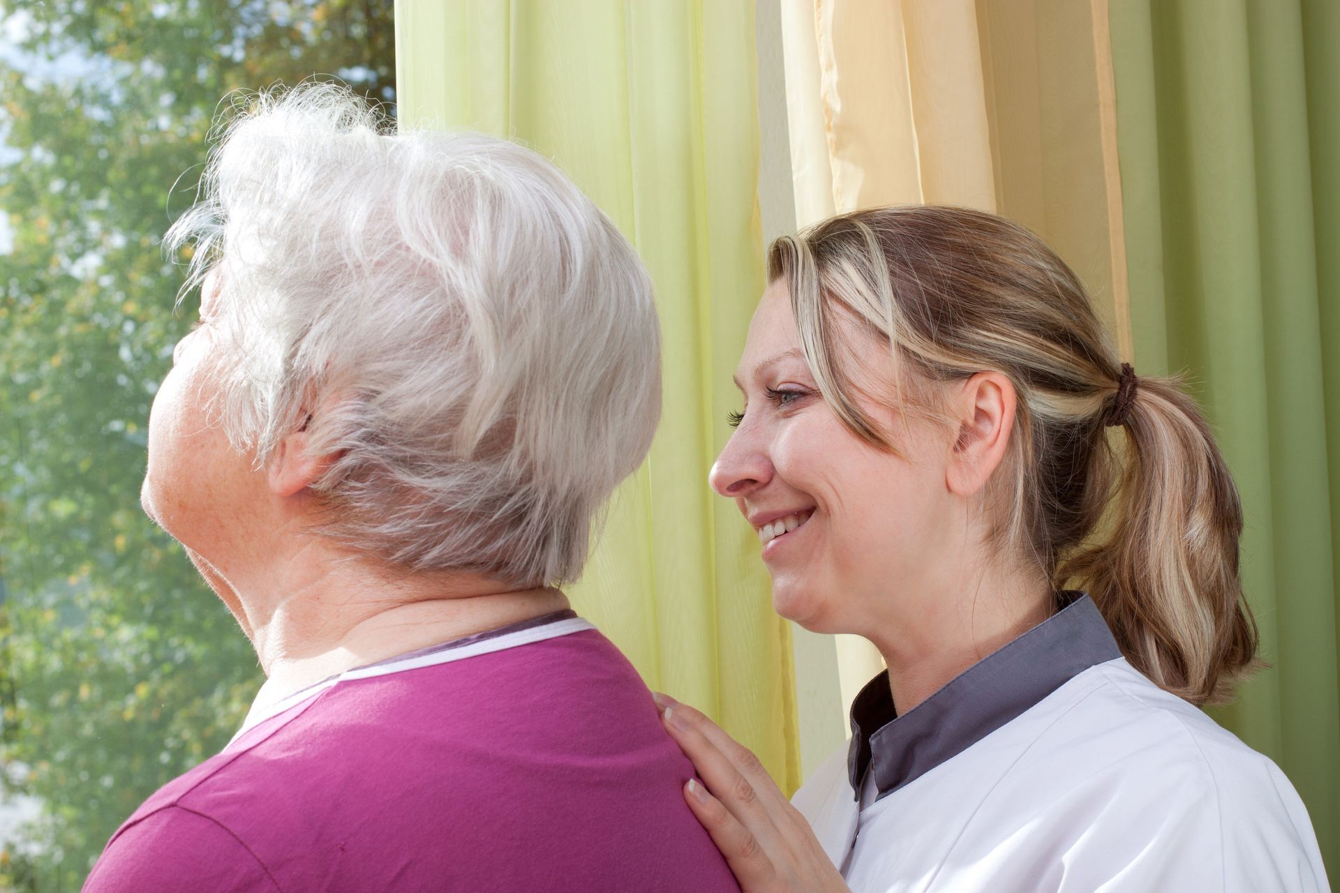 Woman with white hair looks out window as another woman smiles, hand on her shoulder.