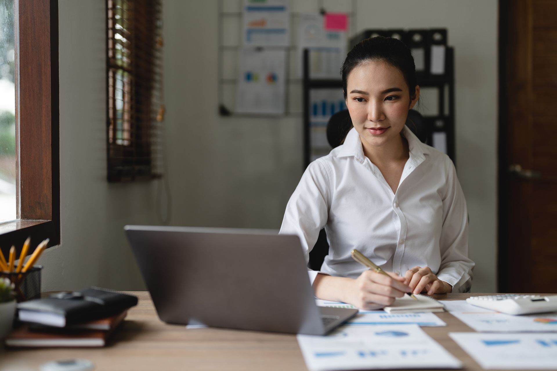 Woman in white shirt, working at desk with laptop, writing in notebook.