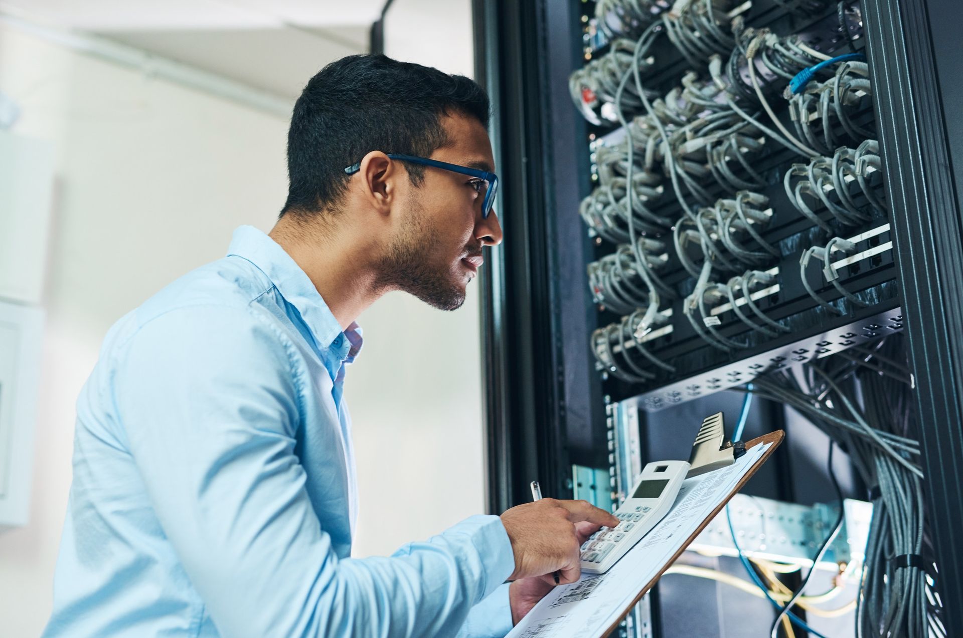 Man inspecting server rack, wearing glasses and blue shirt, holding a clipboard, indoors.