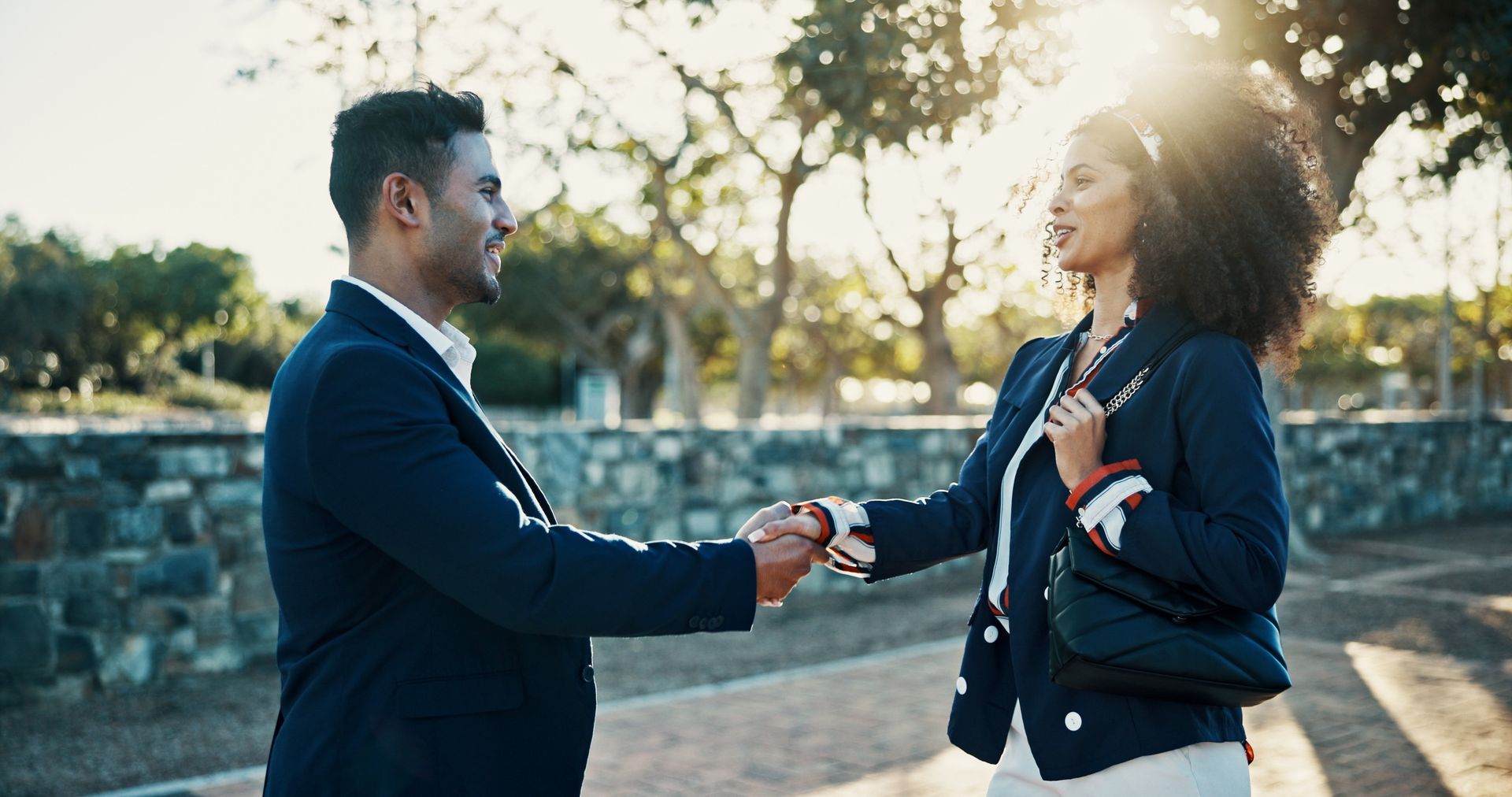 Man and woman shaking hands outdoors, sunny park setting.