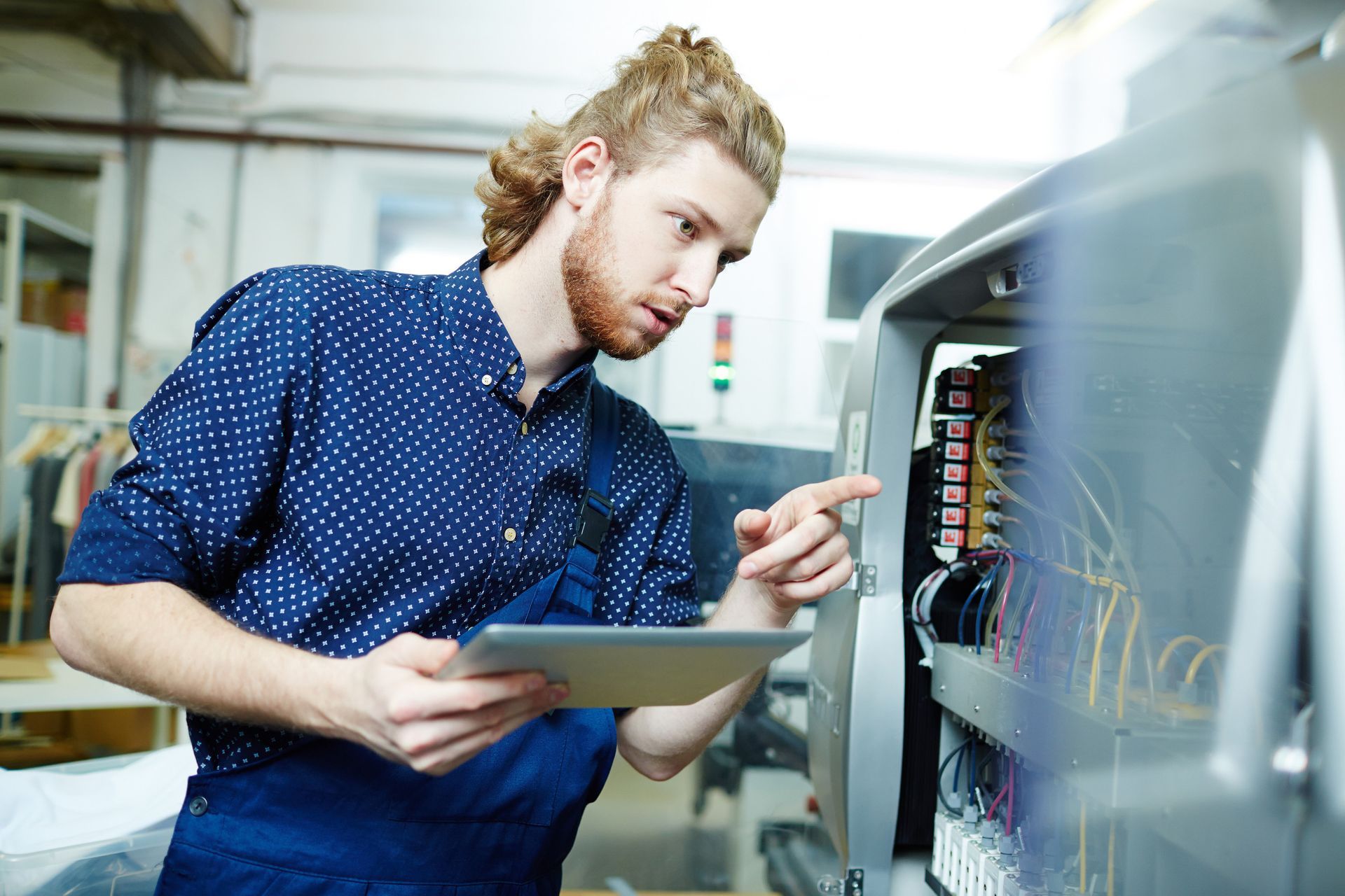 Mechanic examining electrical panel, using a tablet. Workshop setting.