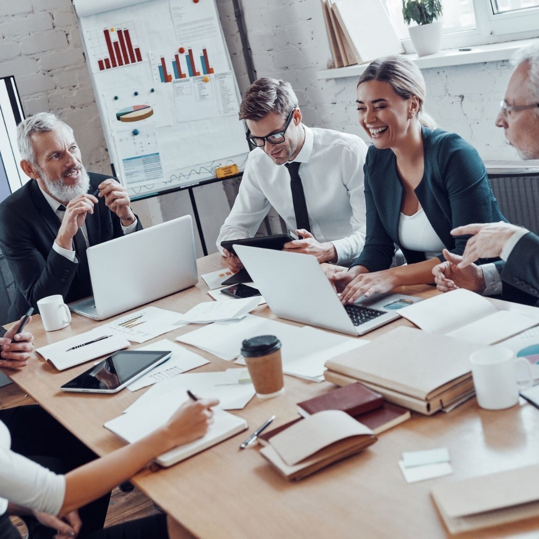 Business team at a conference table with laptops, papers, and a whiteboard, discussing data and smiling.