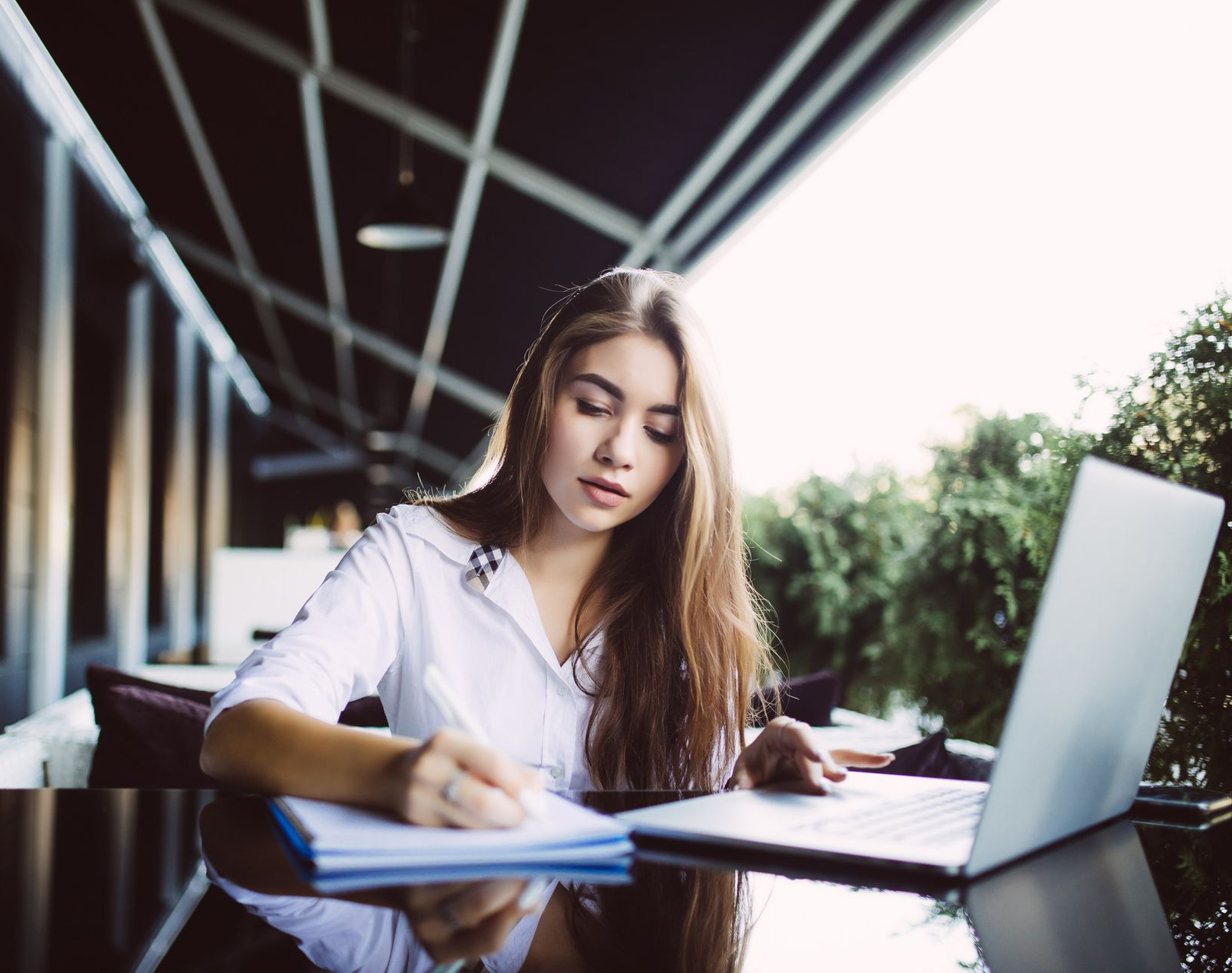 Woman writing in notebook, using laptop outside at table; sunlight.