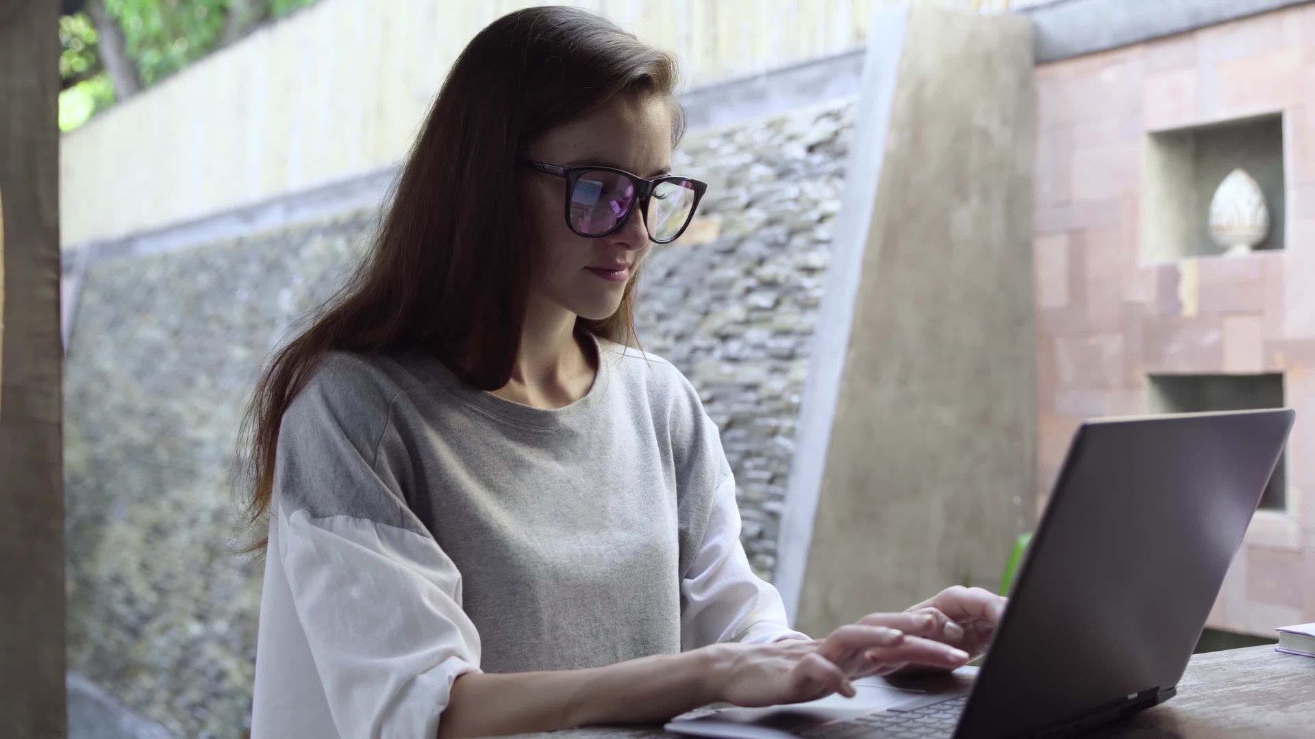 Woman with glasses working on a laptop outdoors, gray and white top, neutral background.