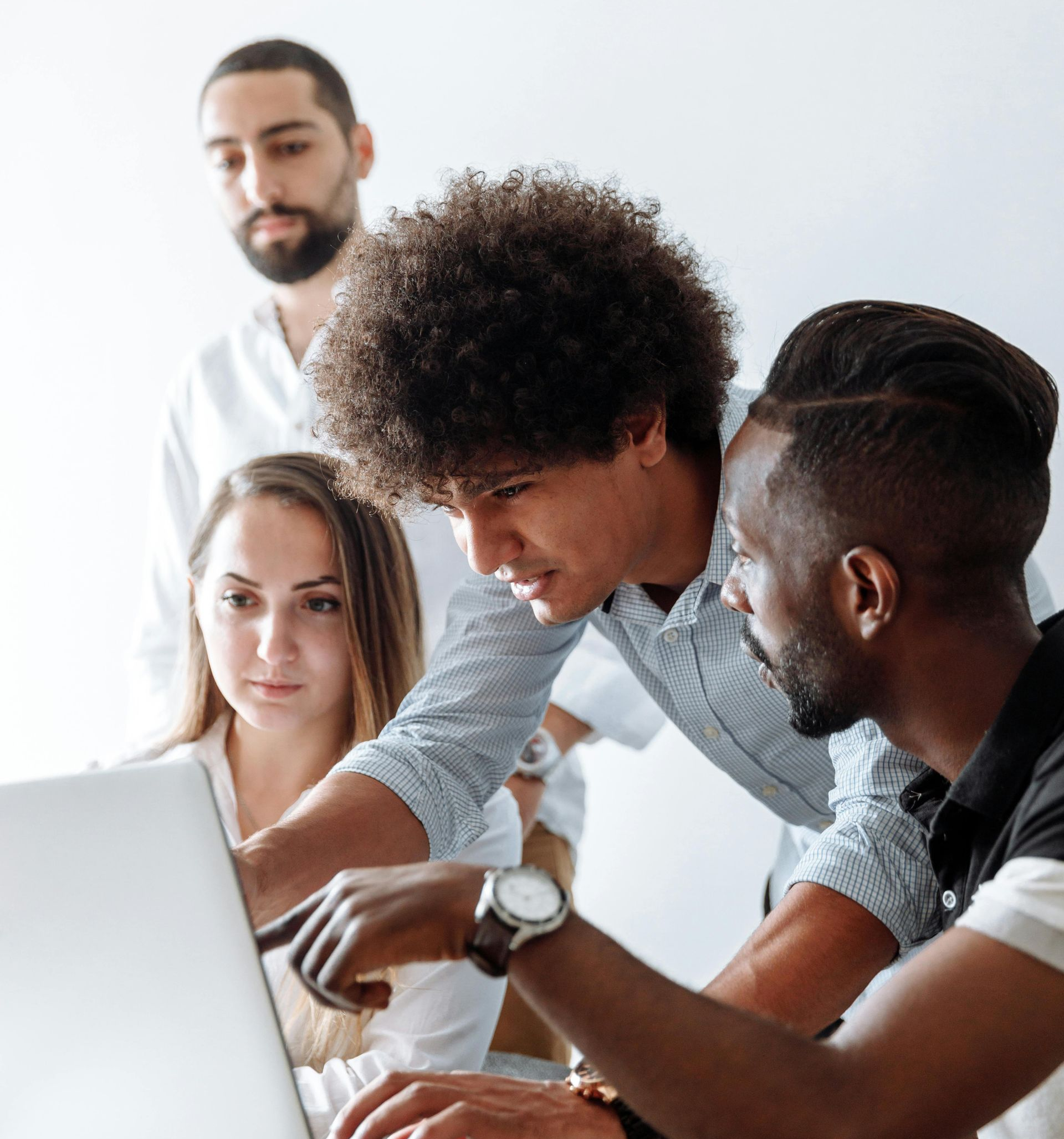 Four people looking at a laptop, pointing at the screen. Bright room, focused expressions.