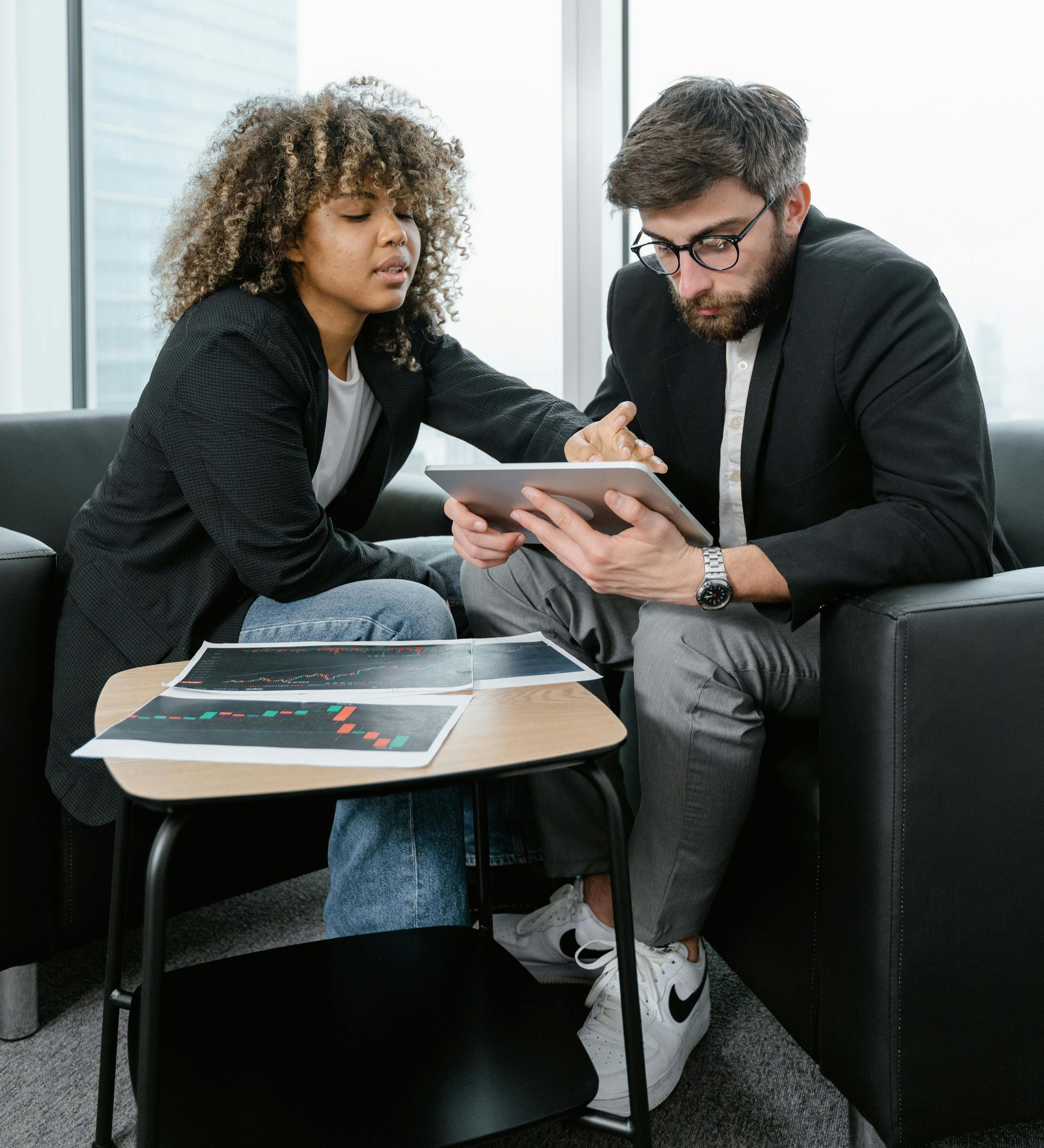 Woman and man in business attire review documents on a tablet; seated on a sofa.