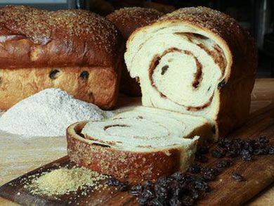 A loaf of bread is sitting on a wooden cutting board next to raisins and flour.