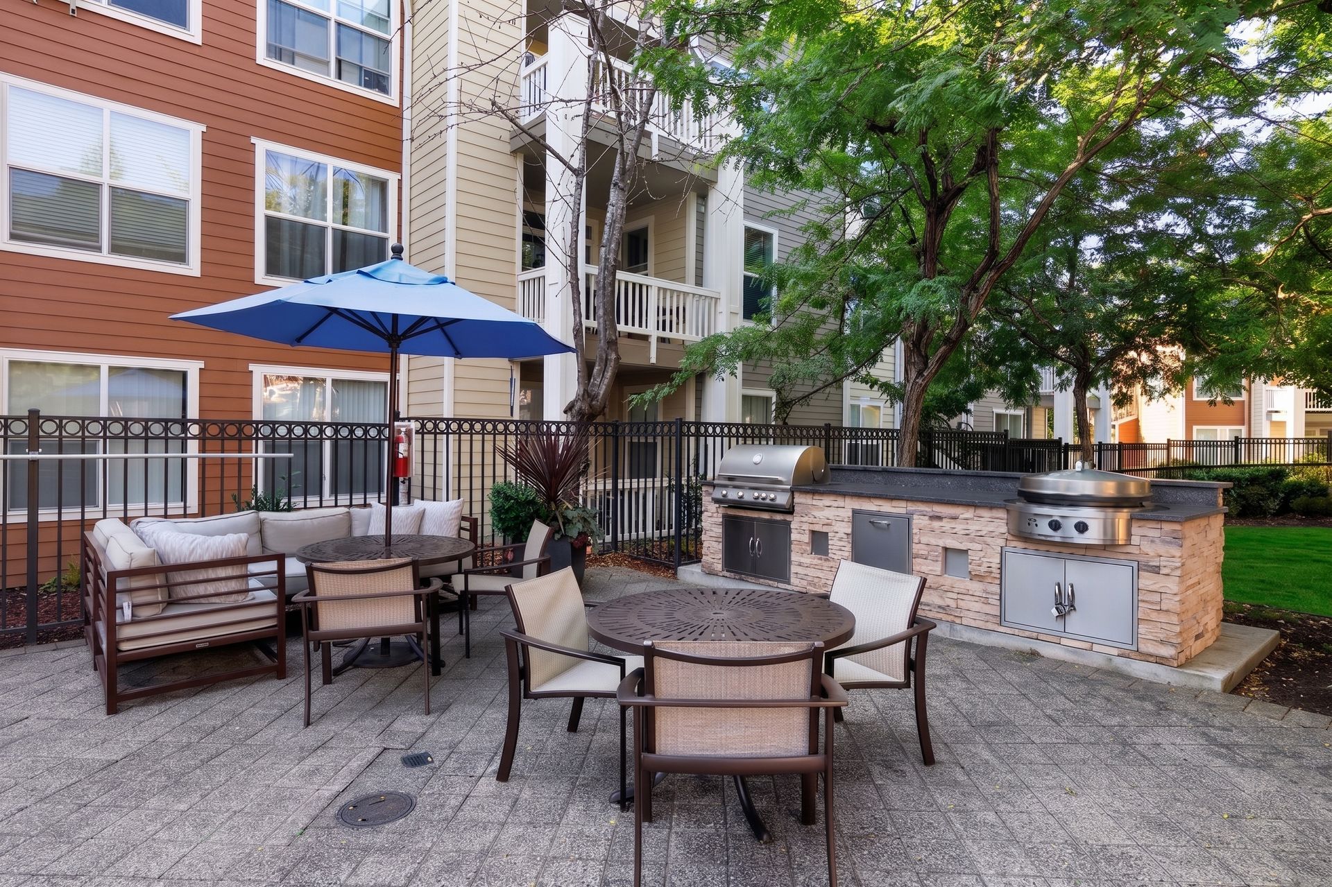Outdoor patio with tables, chairs, umbrellas, and a brick grill area beside an apartment building.