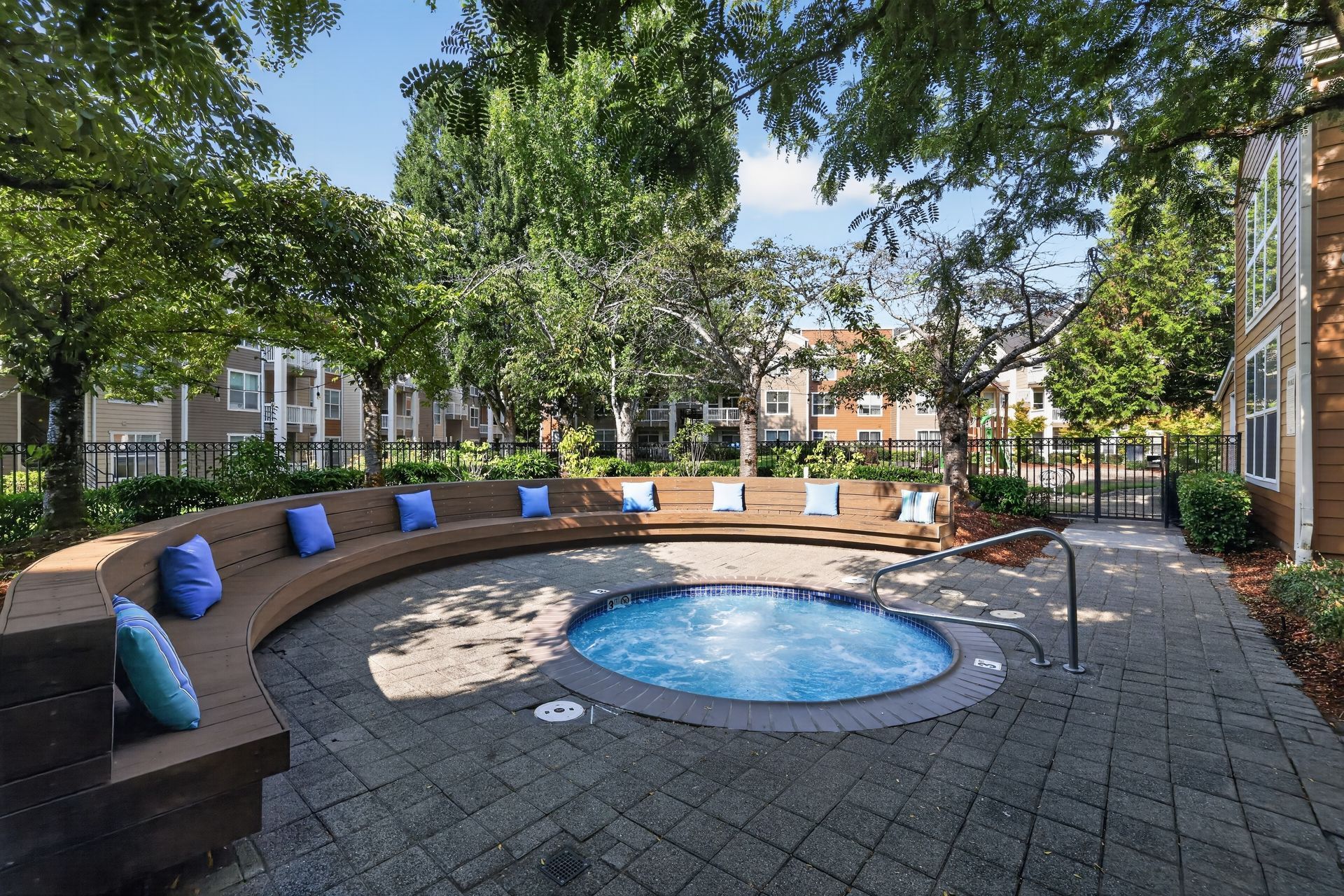 Outdoor courtyard with a circular hot tub, curved bench seating, and blue cushions under trees